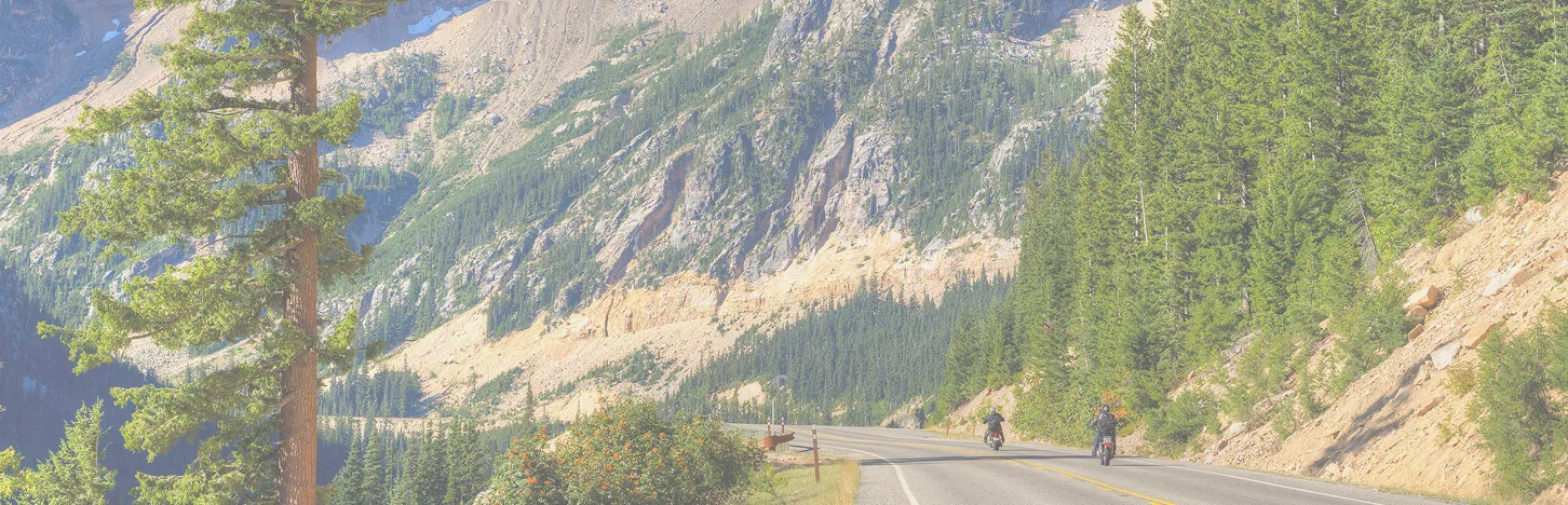Scenic mountain highway through forested cliffs with two motorcycles riding along a winding road in the North Cascades.