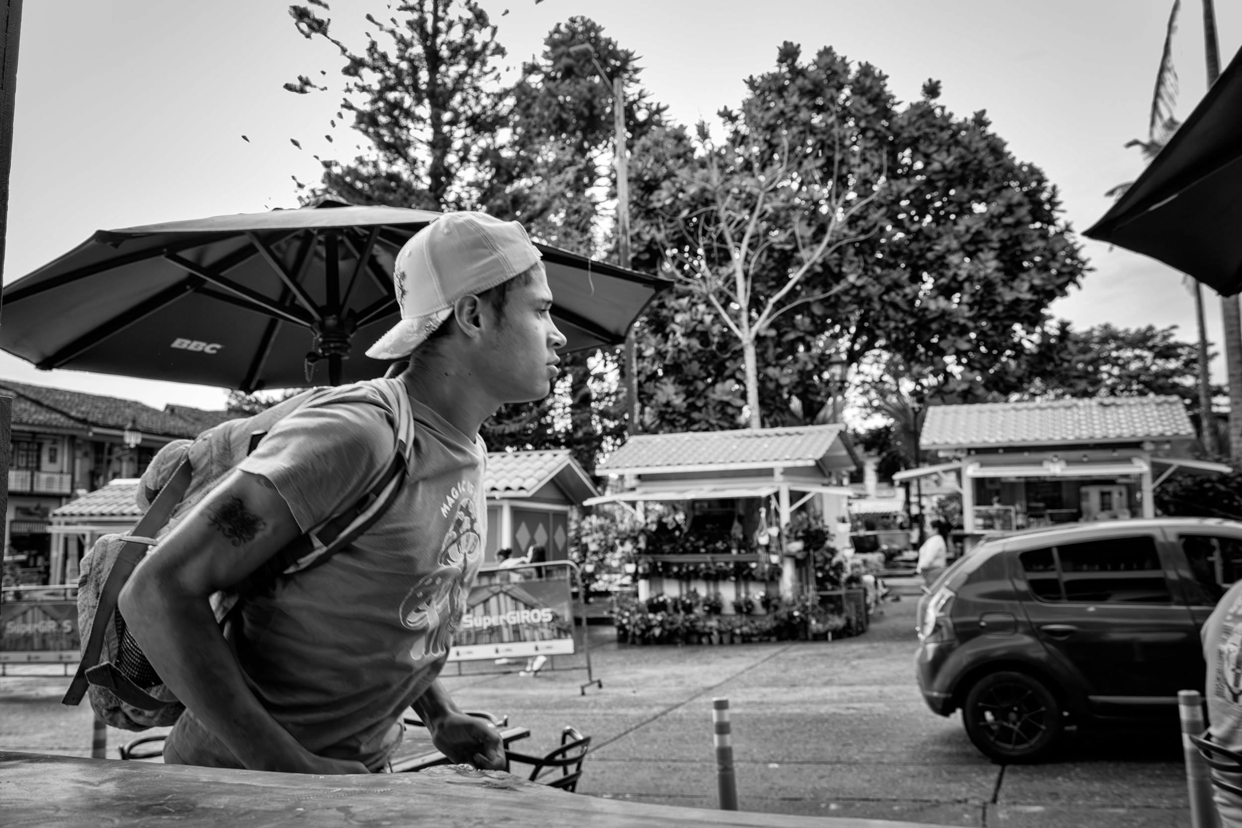 A person in a hat leans over a table outdoors, surrounded by trees and parked cars in a black-and-white setting.