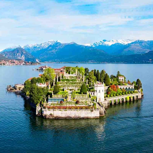Lush, terraced garden on a small island surrounded by a lake, backed by snow-capped mountains under a partly cloudy sky.