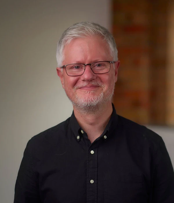 Phot of adam Coles who has grey her and wearing glasses and a black shirt while smiling at the camera