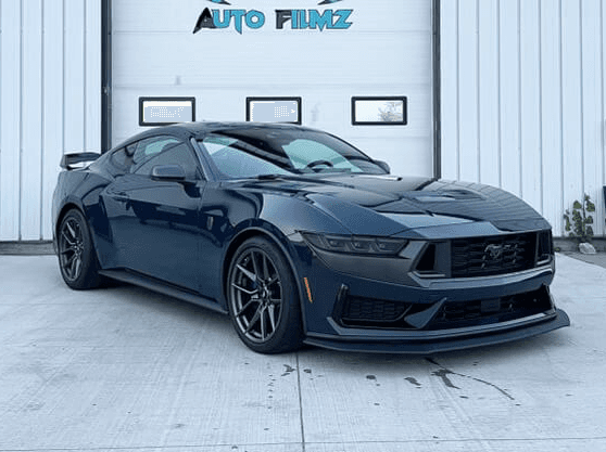 A dark blue Ford Mustang Dark Horse is parked on a concrete driveway in front of a white industrial building.