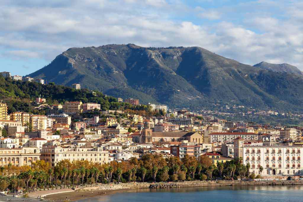 A red gondola car travels on cables over a green valley with a large lake and a town on its shore.