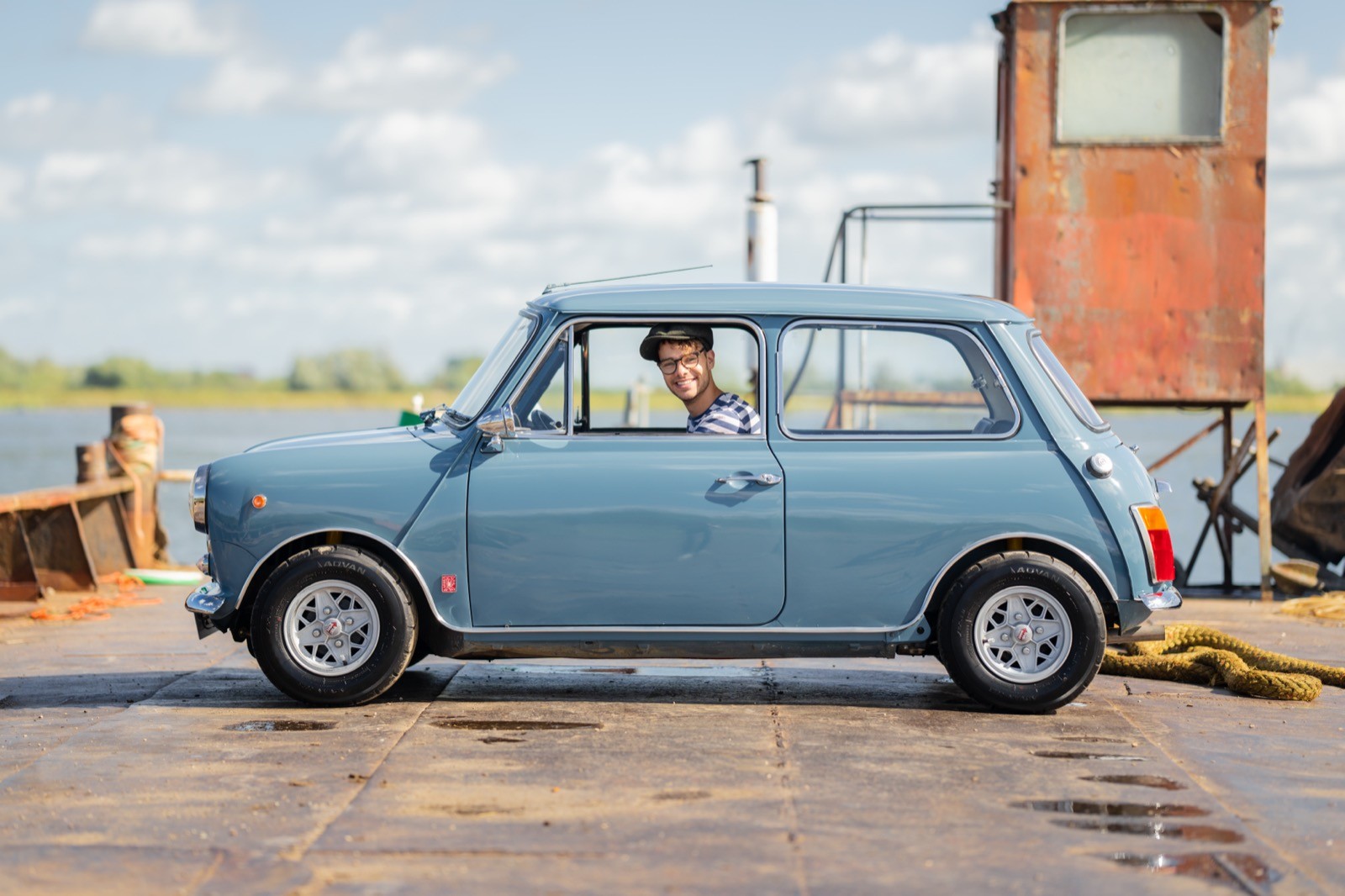 A vintage blue car parked next to a small building by the water under a partly cloudy sky.
