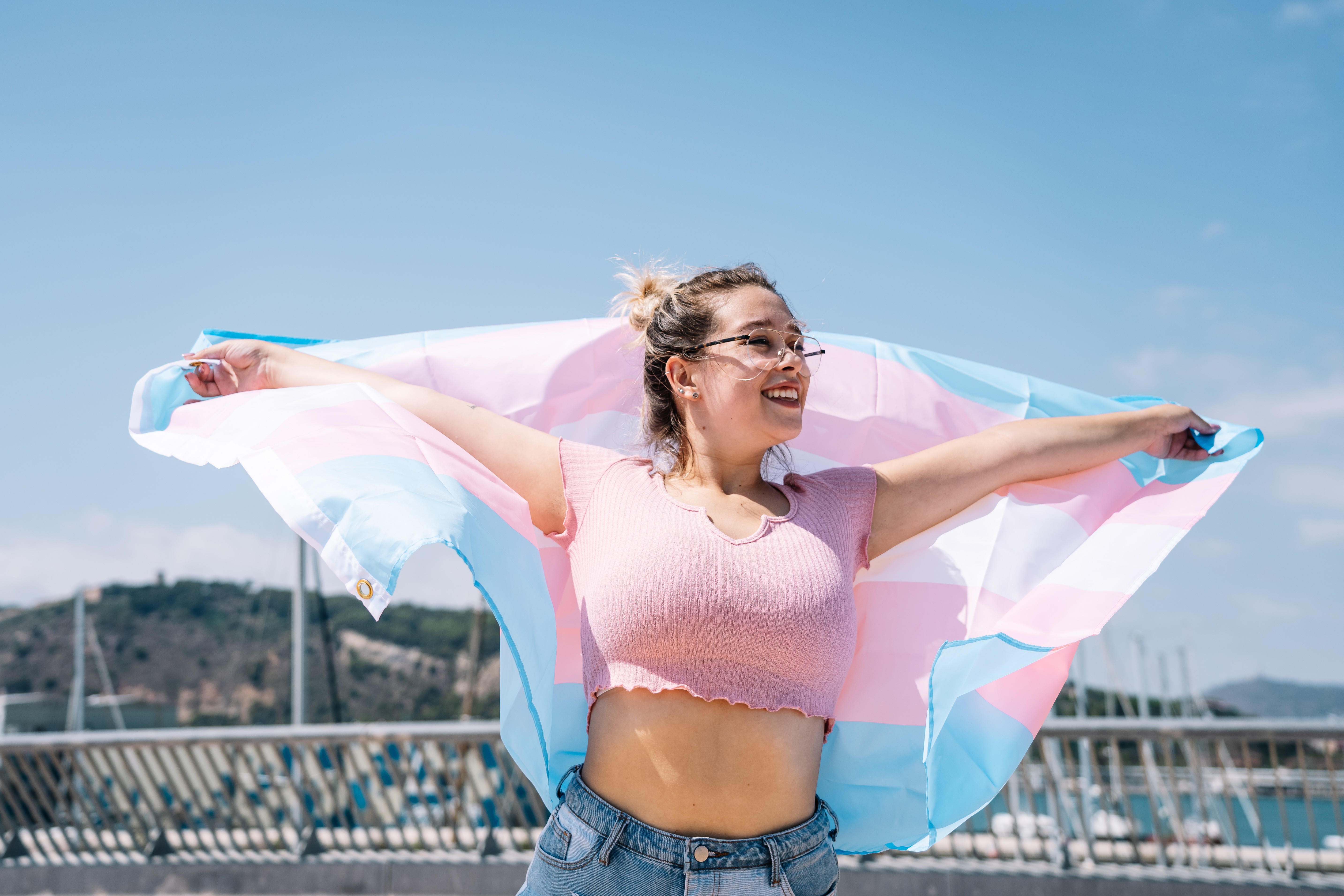 Foto de uma pessoa sorridente ao ar livre, segurando a bandeira trans estendida atrás de si. Ela usa uma blusa rosa e jeans, com o cabelo preso, em um dia ensolarado com céu azul e elementos de uma marina ao fundo.