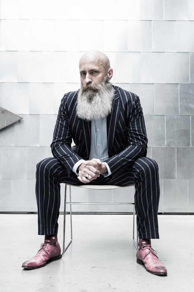 Bas Stronks, marketeer met grote grijze baard en streepjespak en roze scheonen zittend in een industriele omgeving.