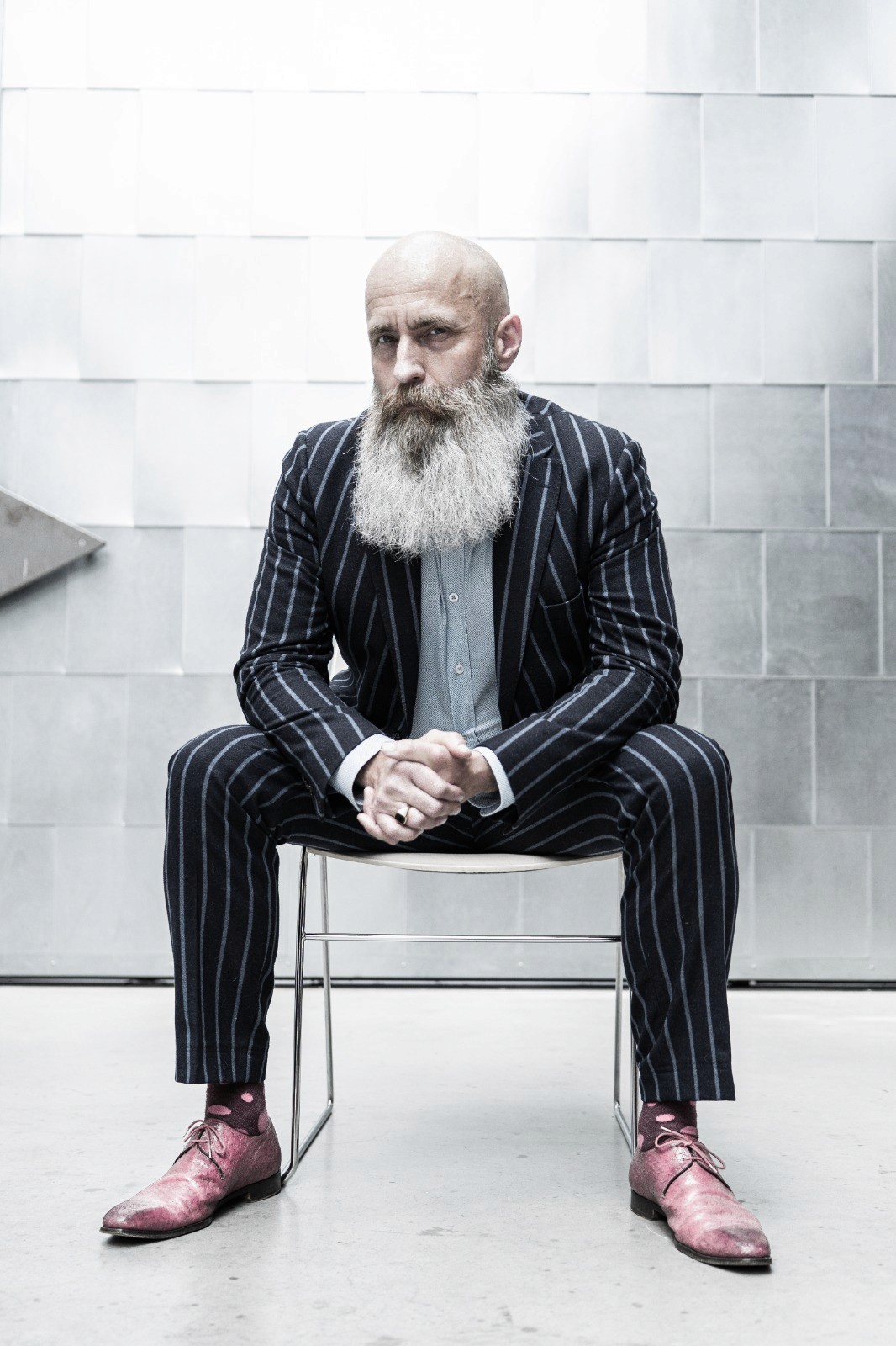 Bas Stronks, man met grijze baard en streepjespak en roze scheonen zittend in een industriele omgeving.