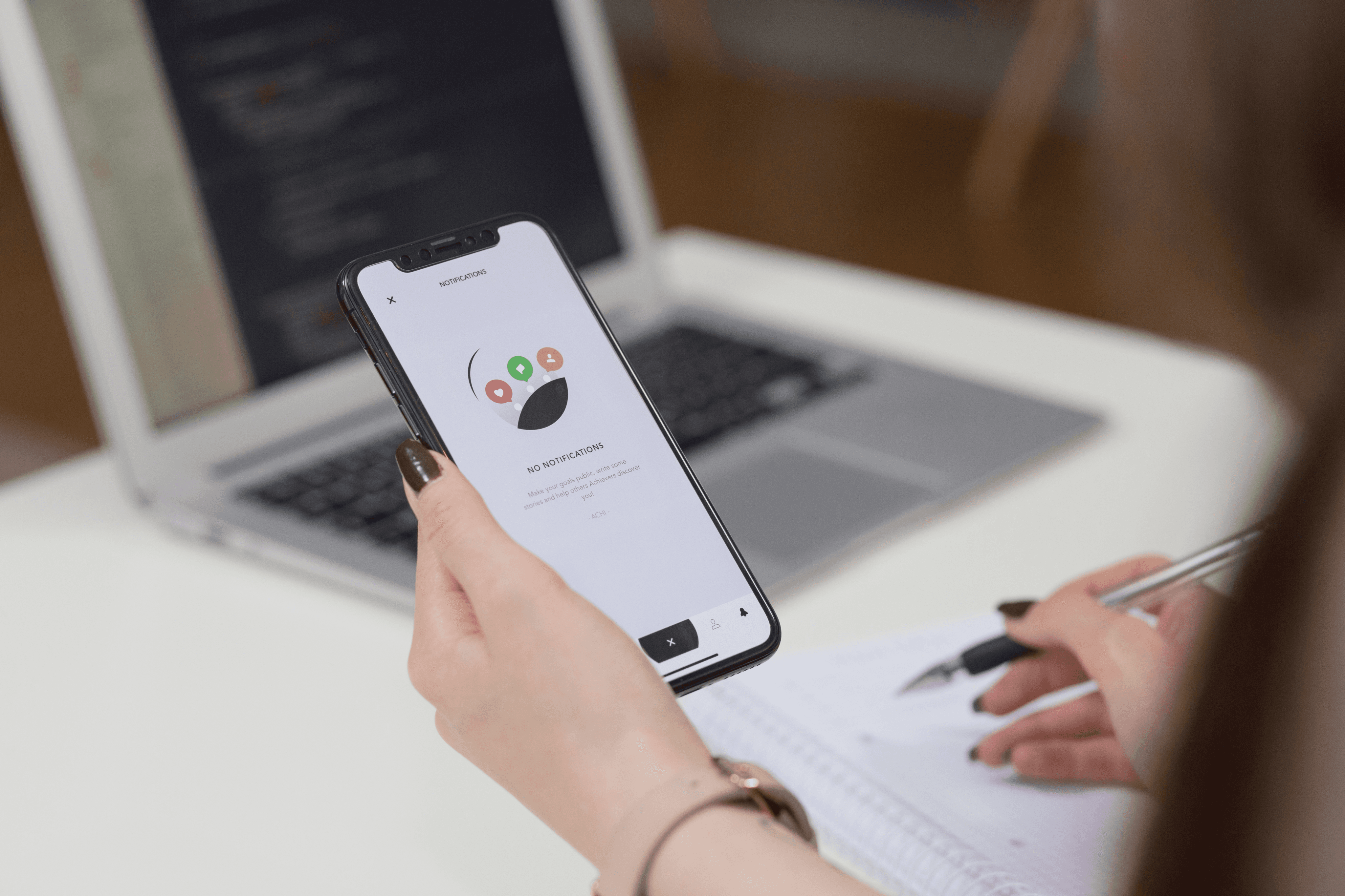 User testing a design prototype on a cell phone and taking notes on a desk with a laptop displaying code in the background - Photo sourced from unsplash by Freestocks.