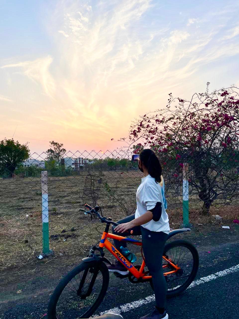 Una persona con una camiseta negra y pantalones de color claro está de pie en la cima rocosa de una montaña al atardecer, sosteniendo una cámara frente a su cara para tomar una foto. Un mar de nubes se extiende detrás de ellos, iluminado por la luz dorada del sol poniente.