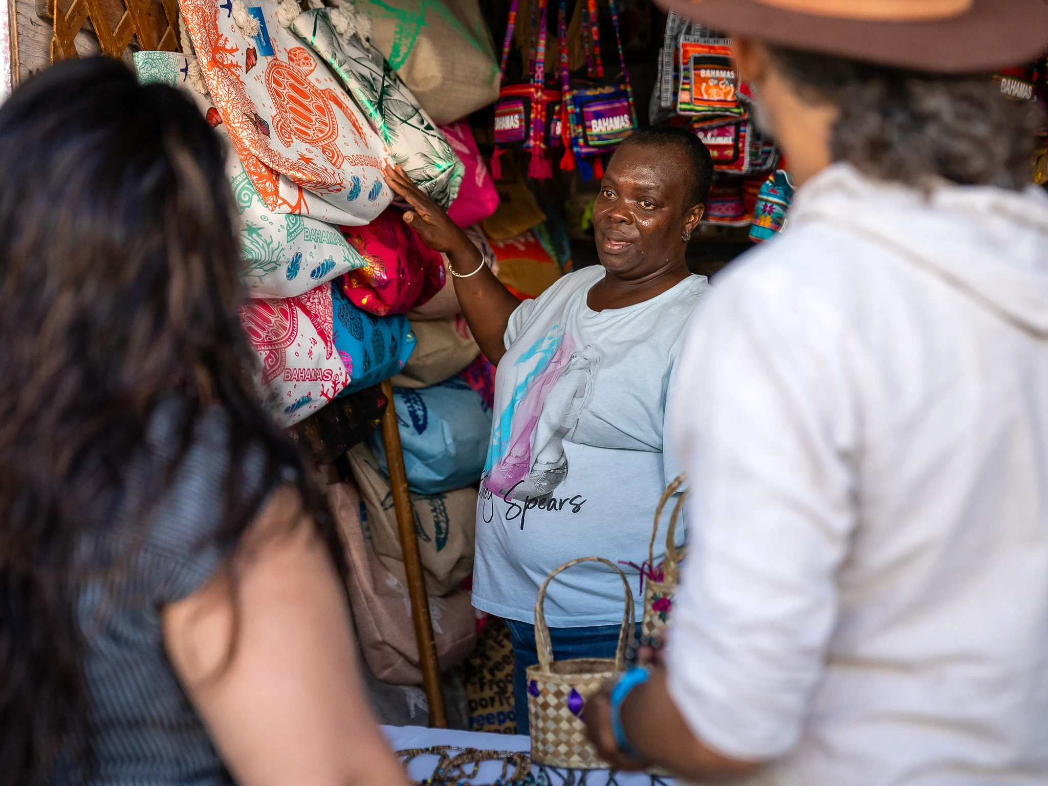 A market vendor smiles while showing colorful bags to two customers in a small stall. The vendor is standing among vibrant textiles, and one customer is holding a basket. The setting is lively and filled with various handmade items.