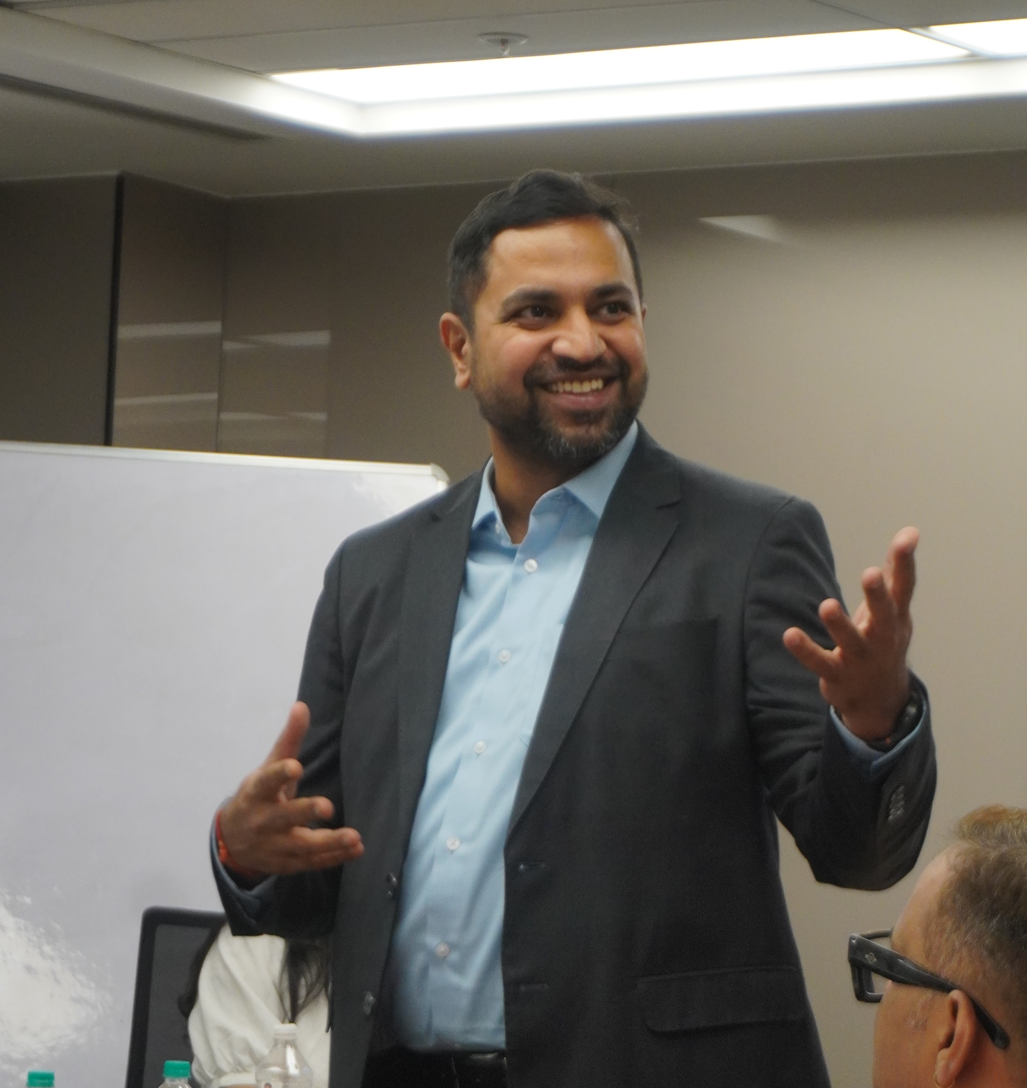 Confident man standing in a greenhouse surrounded by lush green plants, wearing a beige shirt and neutral-toned outfit.