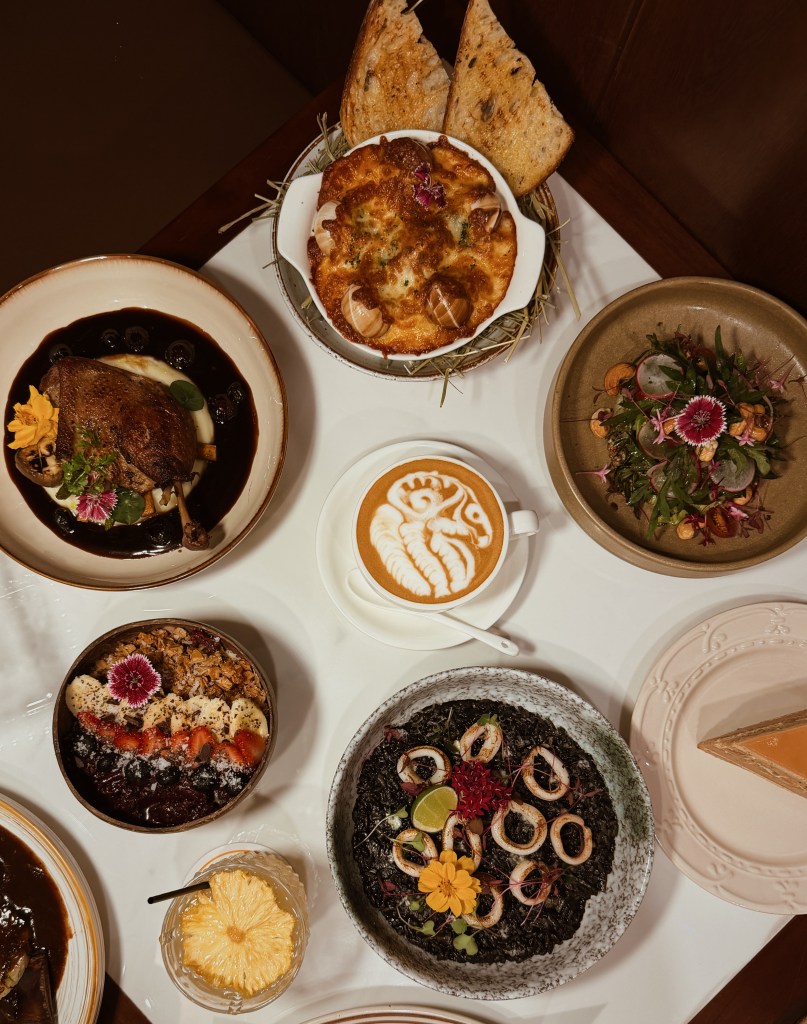 An overhead view of a gourmet meal featuring various dishes including roasted duck with sauce, a baked dish with cheese, a latte with decorative foam, a colorful dessert bowl with fruits, a black rice dish topped with seafood, a cocktail with pineapple, and a slice of dessert on a plate.