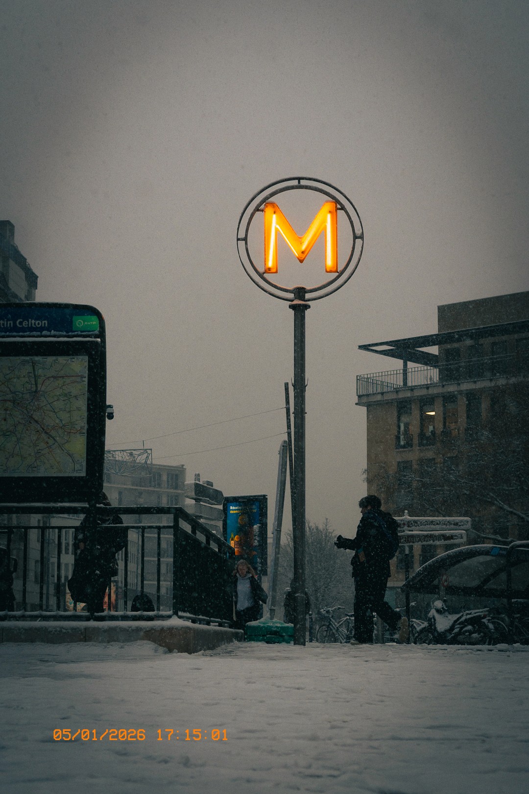 Panneau lumineux M illuminé dans une rue enneigée à Issy, avec des personnes marchant sous la neige, un arrêt de bus et des vélos recouverts de neige. Ciel gris et tempête de neige.