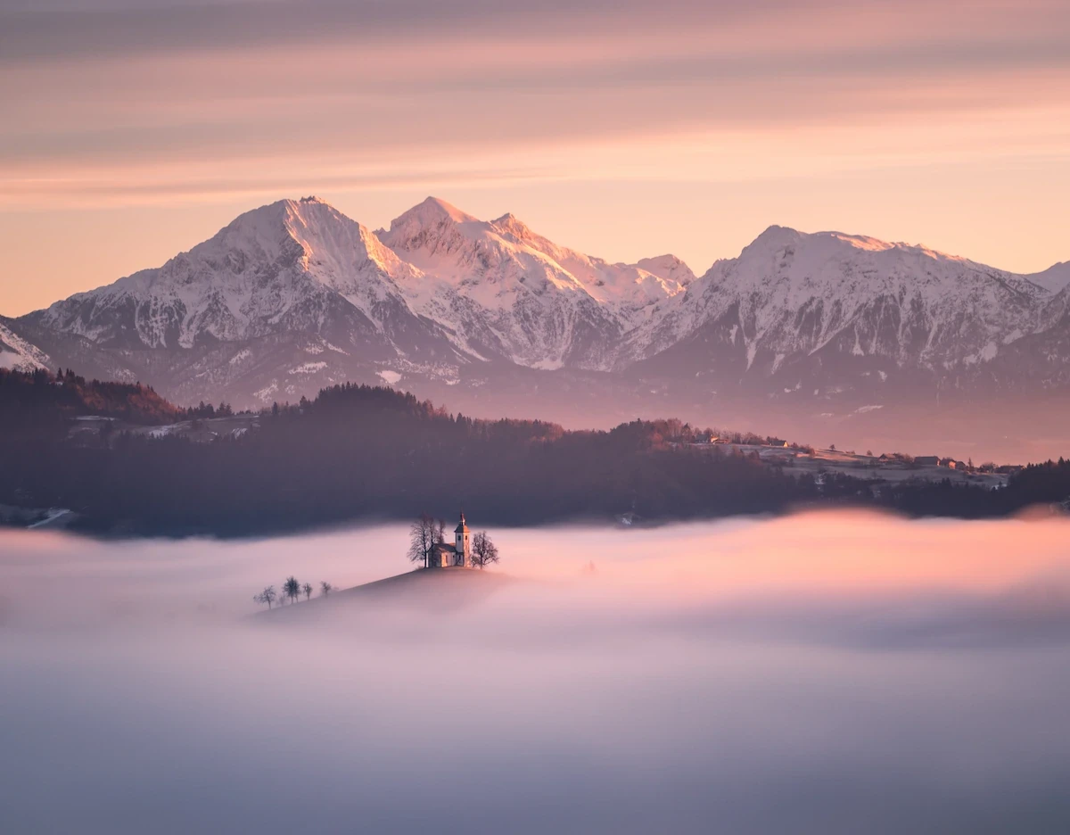 The iconic hilltop church of Sveti Tomaž in Slovenia, appearing as an island above a dense layer of low-lying fog during a pastel sunrise.