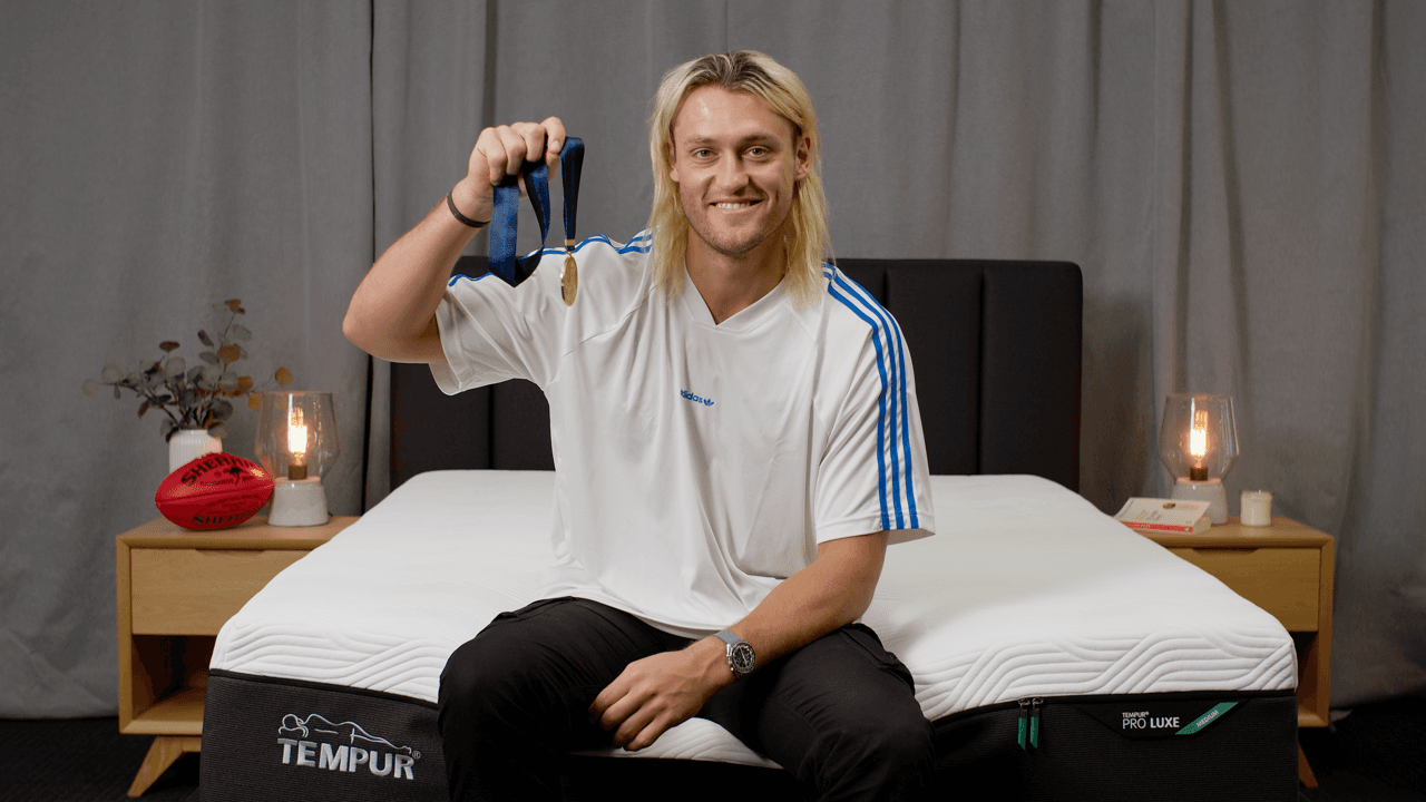A person with blonde hair sits on a bed wearing a light-colored sweatshirt, looking directly at the camera