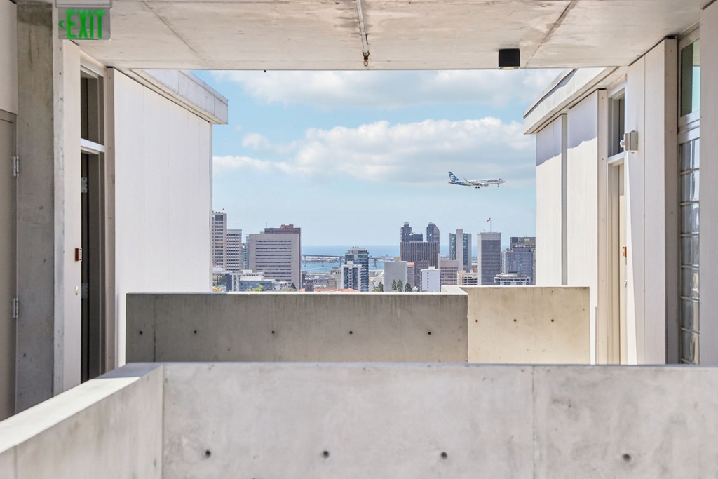 City view through a concrete walkway showing a distant city skyline of San Diego from Bankers Hill, under a blue sky with scattered clouds, a plane mid-flight above tall buildings landing at the airport
