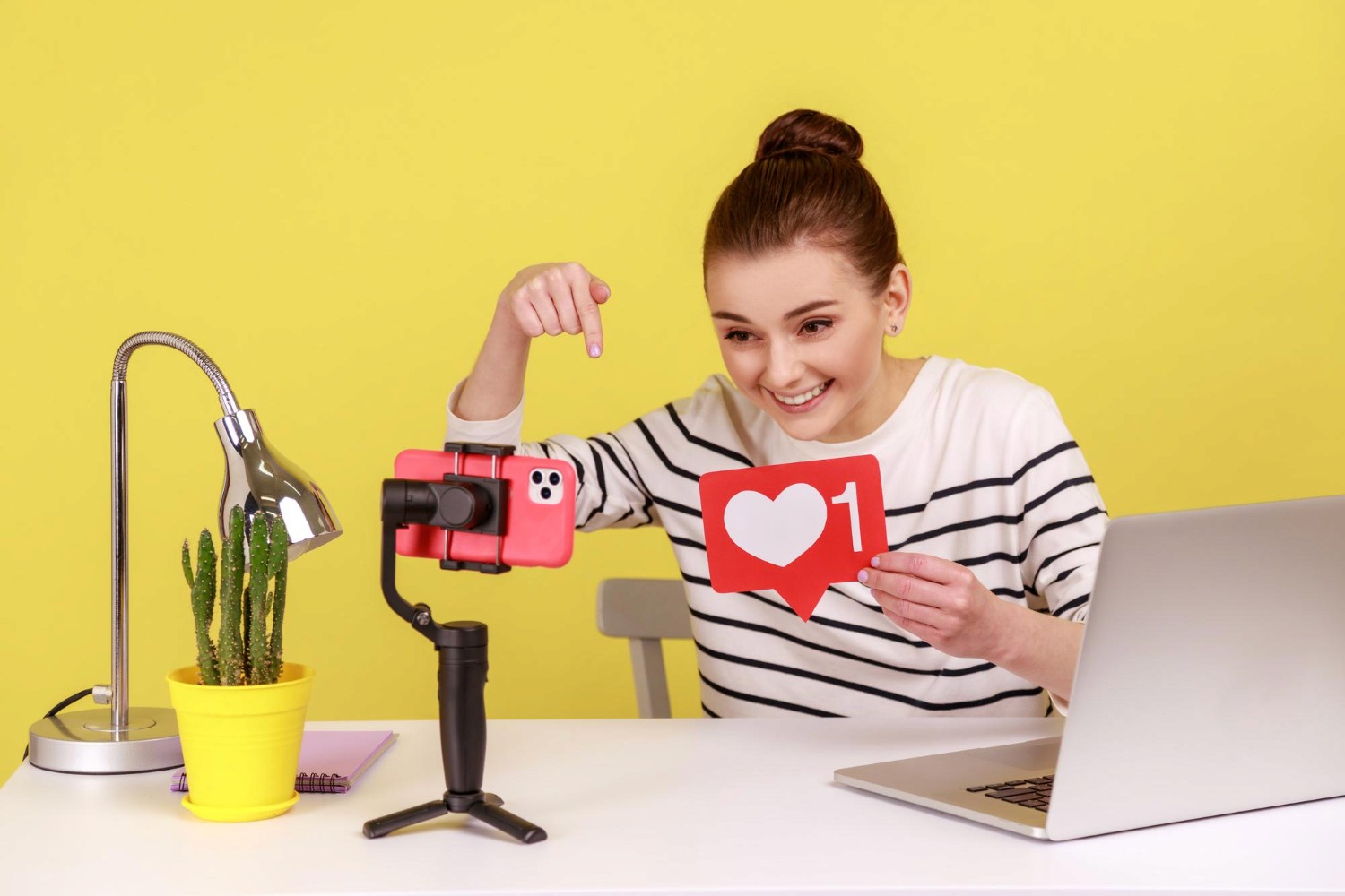 A woman holds a heart-shaped object while sitting in front of a laptop, symbolizing love and technology.