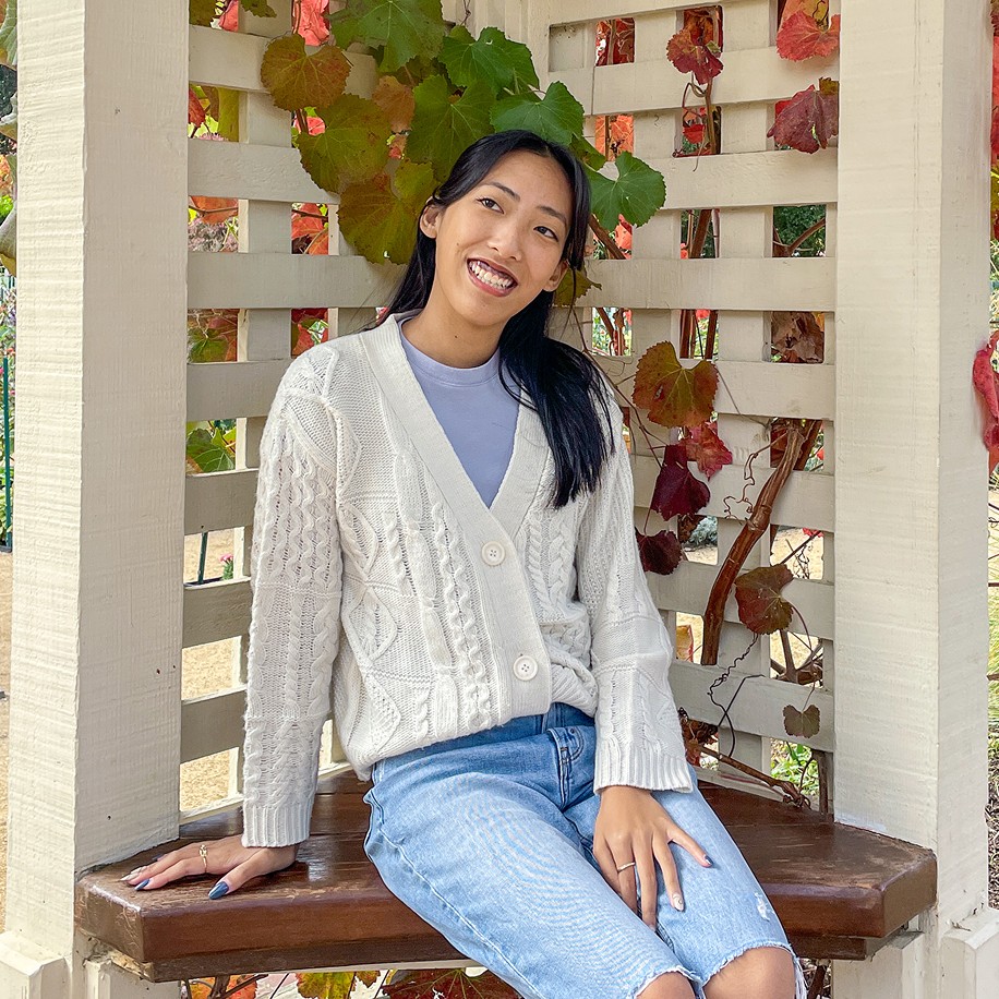 Rebecca's face! She is wearing a white cardigan over a lavender tee shirt and looking off to the left. She's sitting on a bench in front of a trellis.