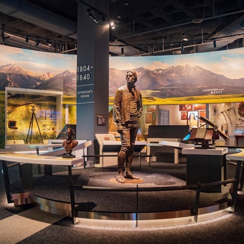 Museum exhibit featuring a bronze statue of a man, display tables, informational panels, and a mural of mountains in the background.