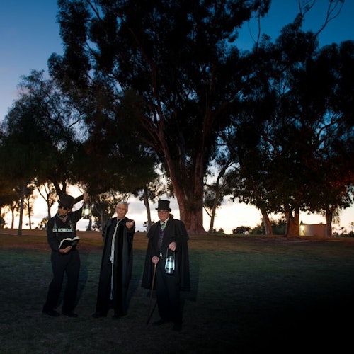 Three people with lanterns and dark clothing stand on grass near trees at dusk. One holds a book and wears a hat labeled "Old Morgue."