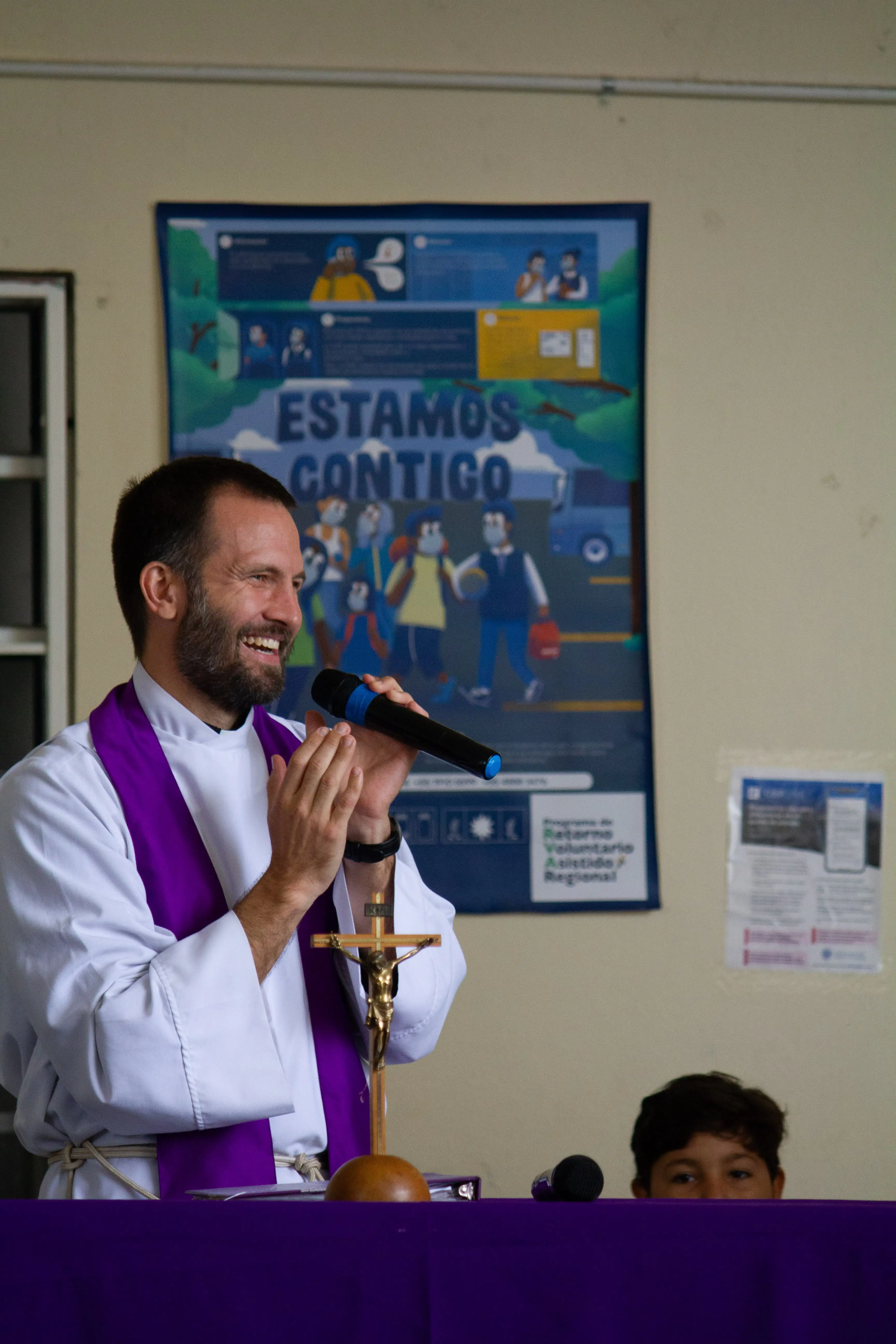A Jesuit priest in religious attire delivers a speech, with a purple stole and a backdrop featuring colorful posters.