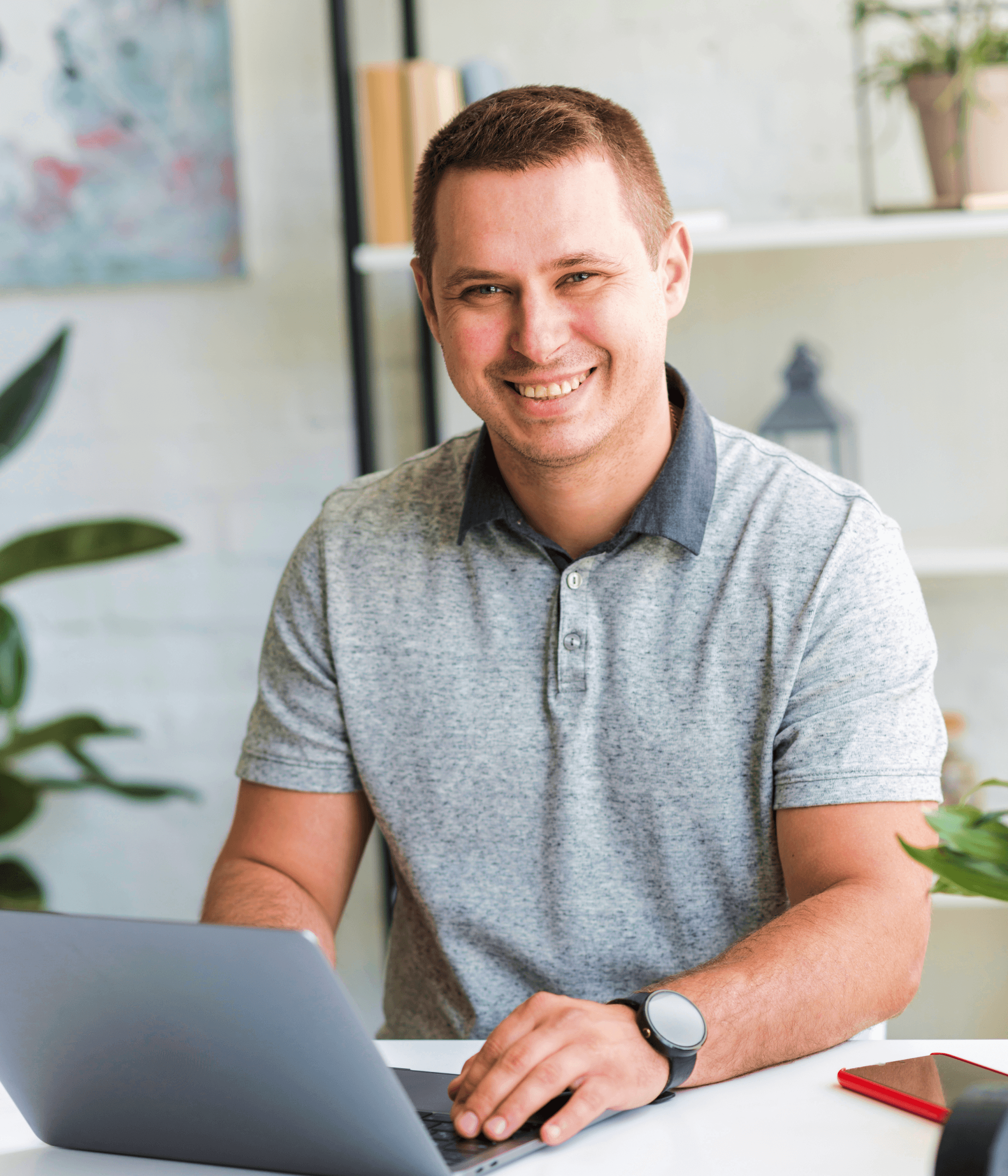 Smiling man wearing a gray polo shirt working on a laptop at a white desk in a bright room with plants.