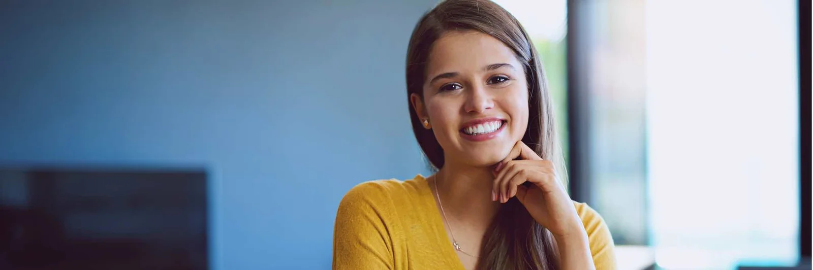 A young woman with long brown hair, wearing a yellow shirt, smiles warmly while resting her chin on her hand in a modern office setting, relating to UCAM Mumbai's welcoming educational environment.