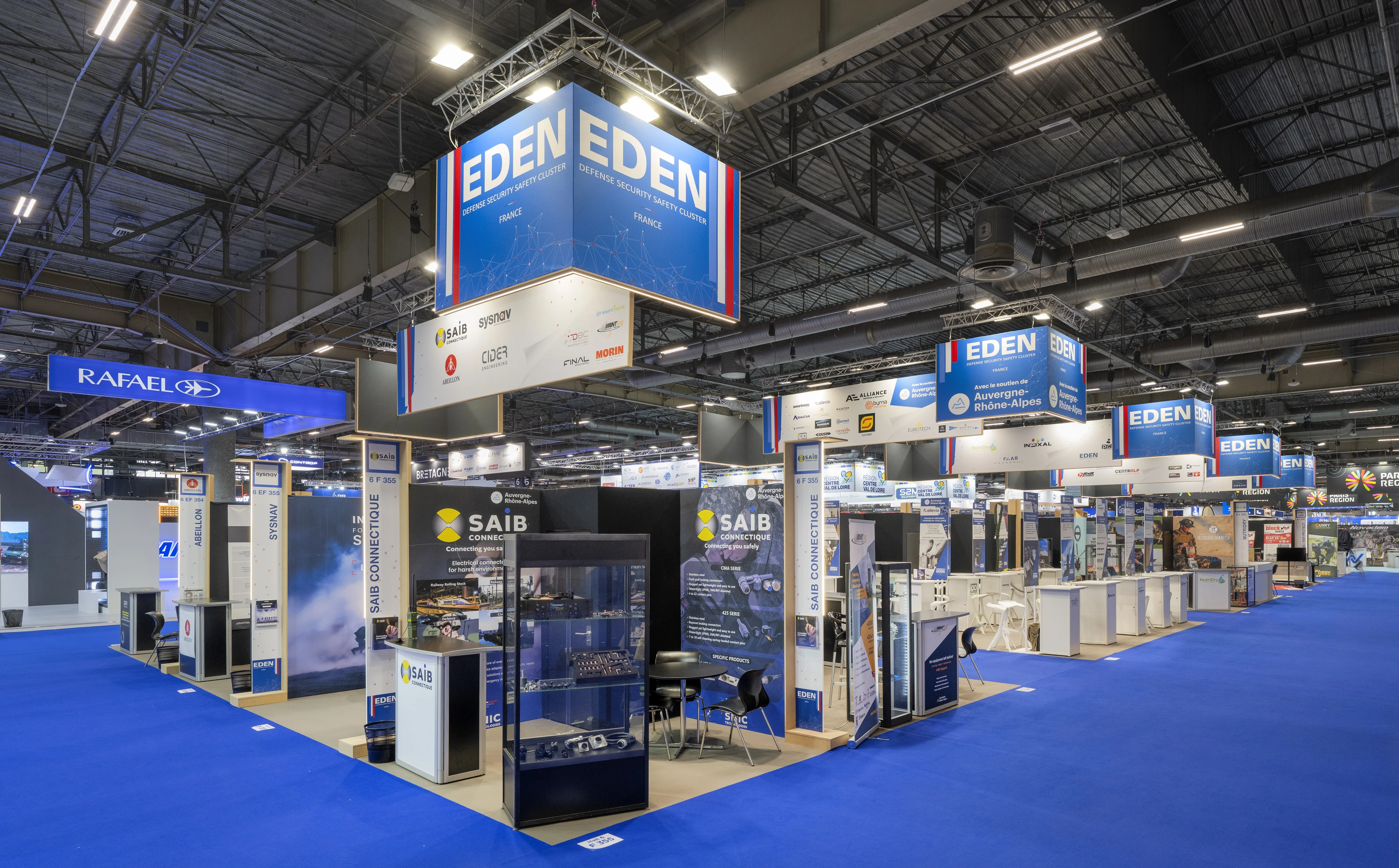 Stand EDEN Cluster conçu et réalisé par Expace à Eurosatory 2026 – pavillon collectif défense bleu-blanc-rouge regroupant les adhérents (SAIB, Sysnav, OBSAM, PGM…) avec vitrines produits, démonstrateurs et accueil central.