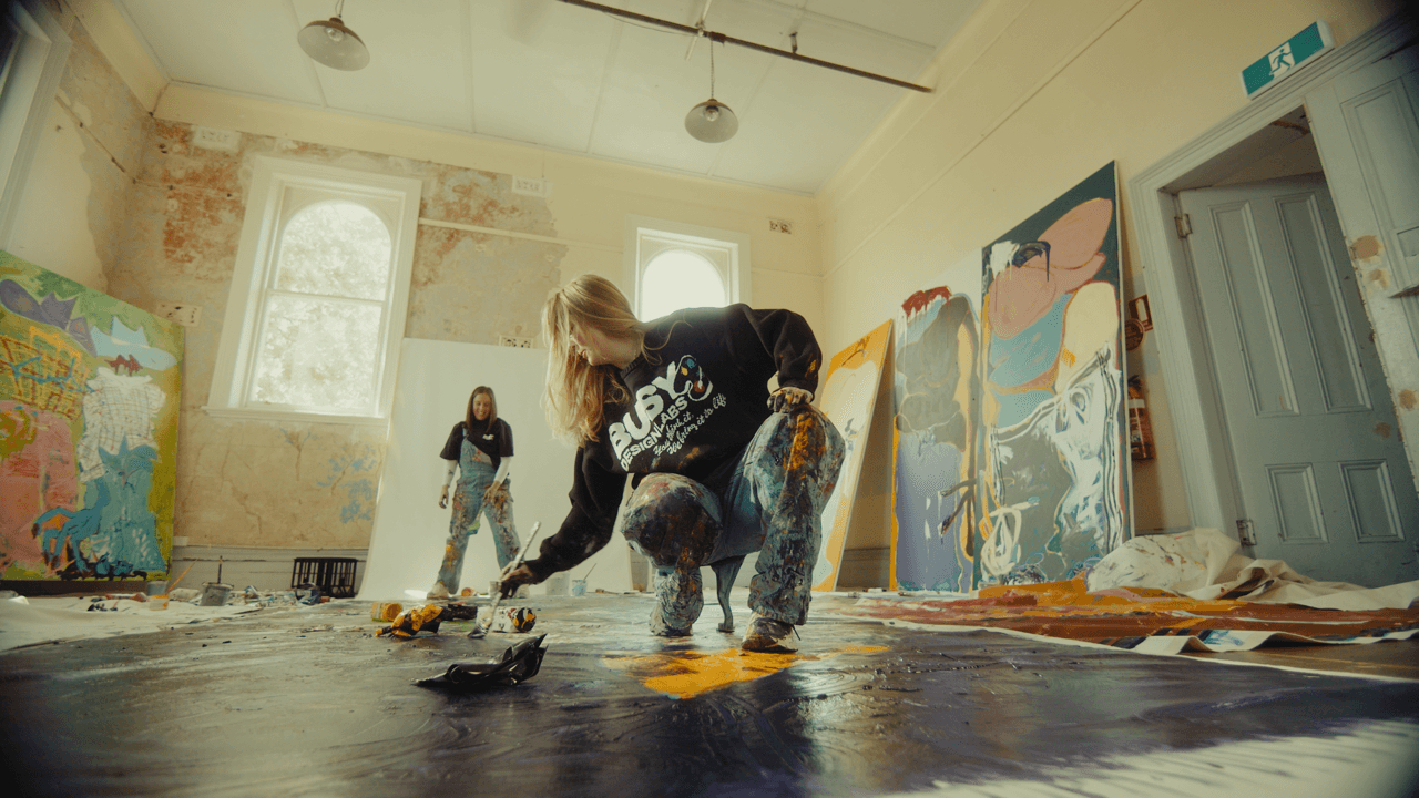 A group of artists working in a spacious studio, surrounded by large canvas paintings and art supplies, creating vibrant, abstract artwork on the floor