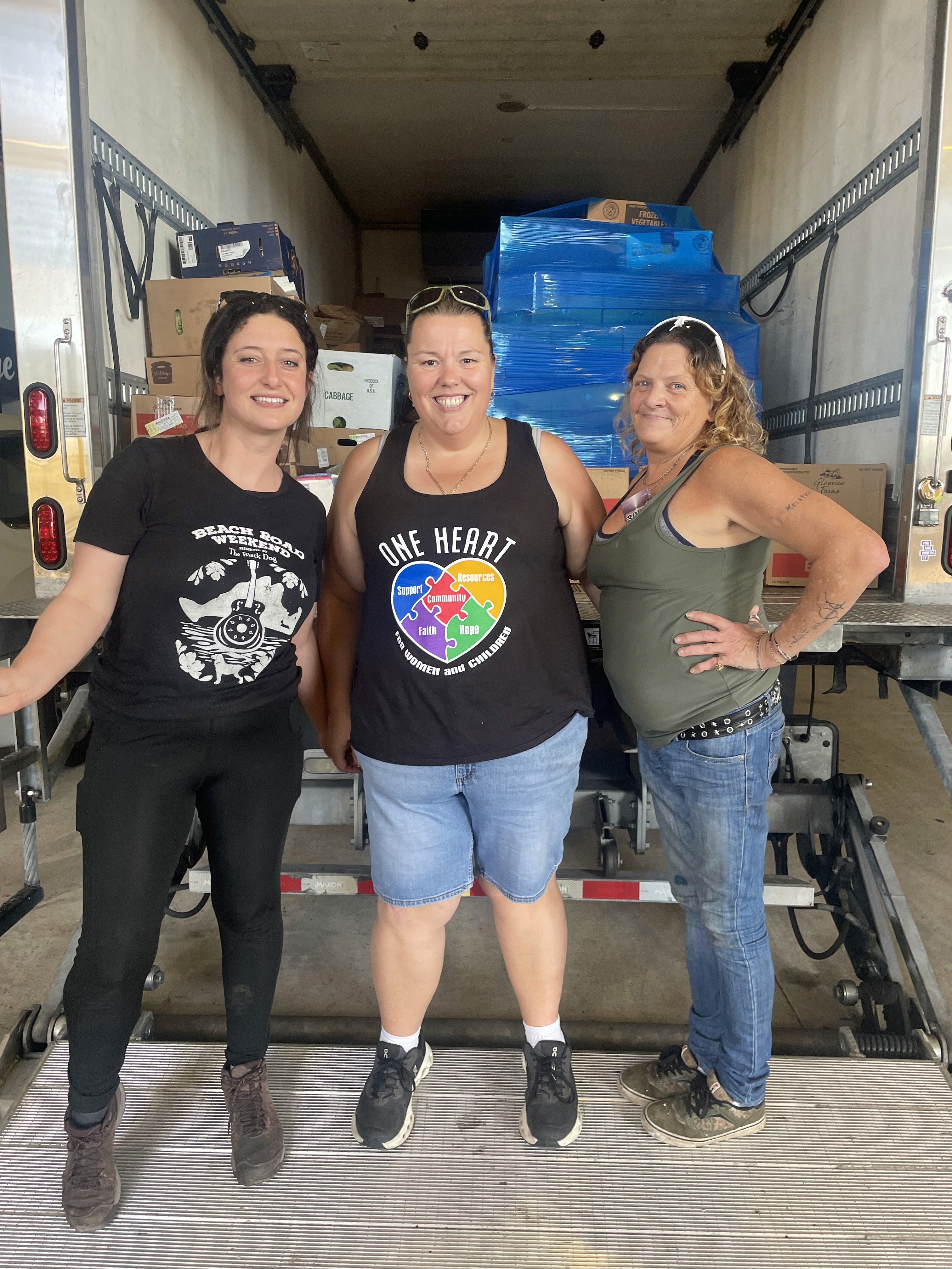 Three smiling women stand together in a truck's loading area, dressed casually and posing for the photo.