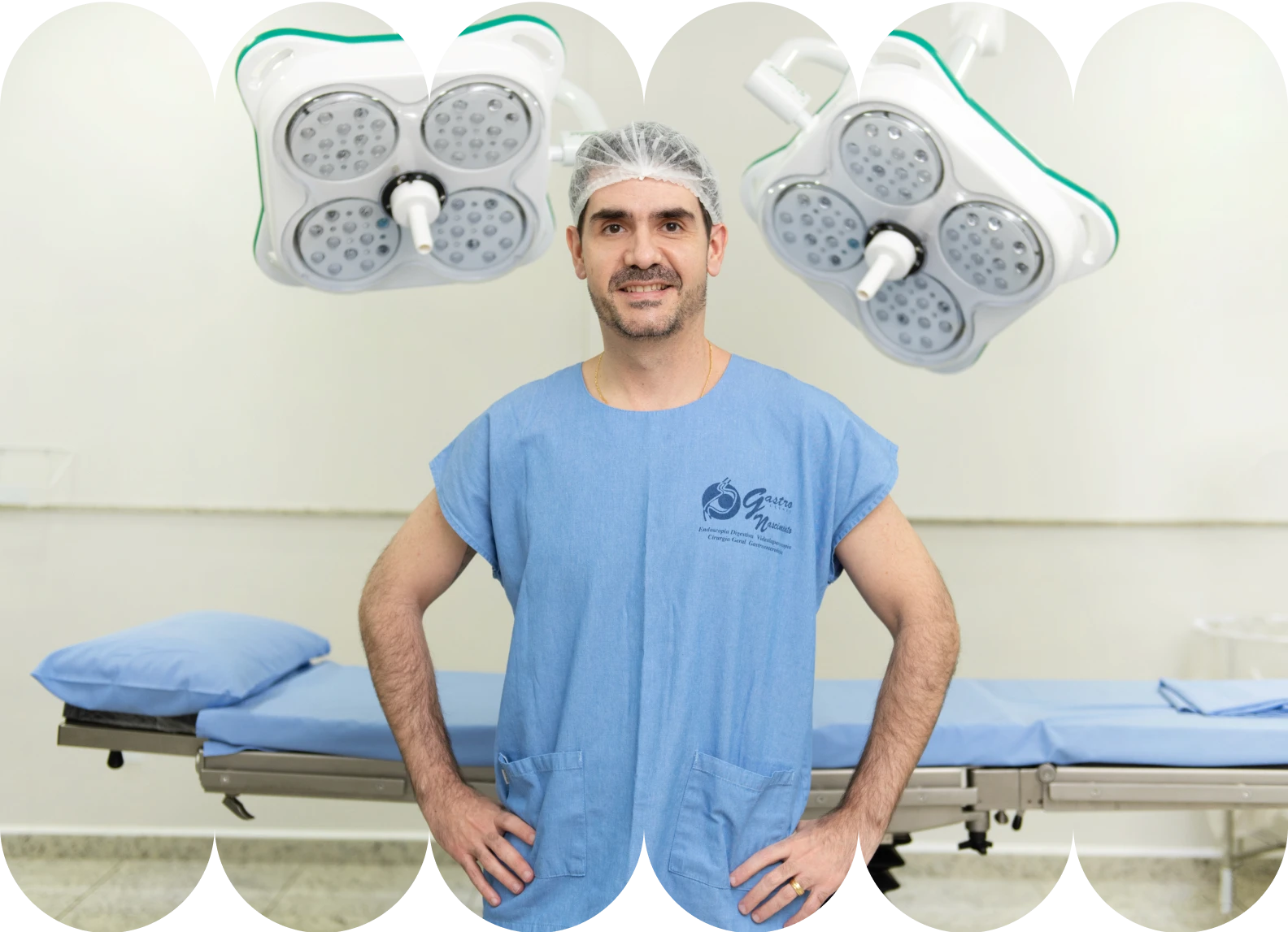 O Dr. Renan sorrindo e olhando para a câmera, vestindo um uniforme cirúrgico azul (scrubs) e touca, em pé em uma sala de cirurgia. Duas luminárias cirúrgicas modernas estão penduradas acima de uma maca cirúrgica.
