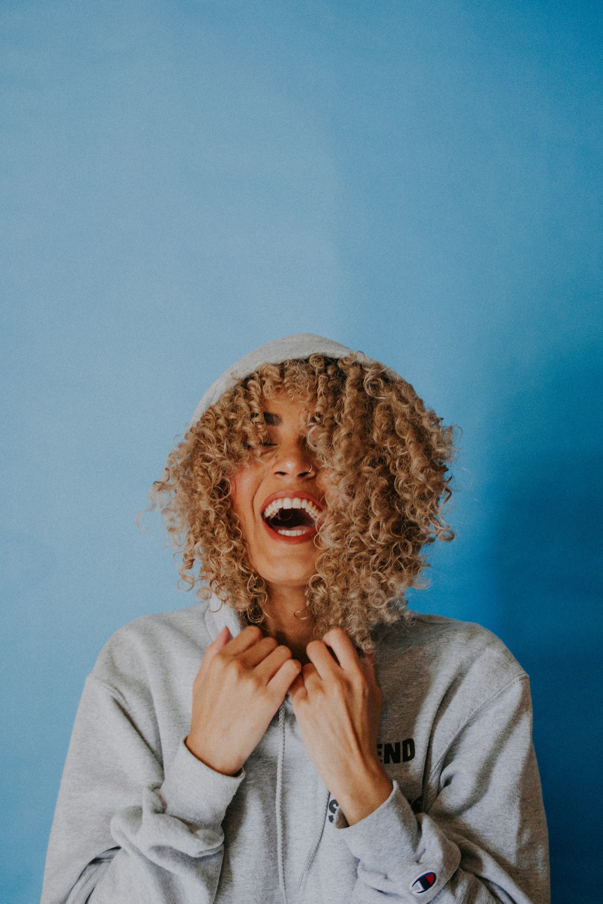 A laughing person with blonde curly hair wearing a grey Champion hoodie, set against a solid blue background.