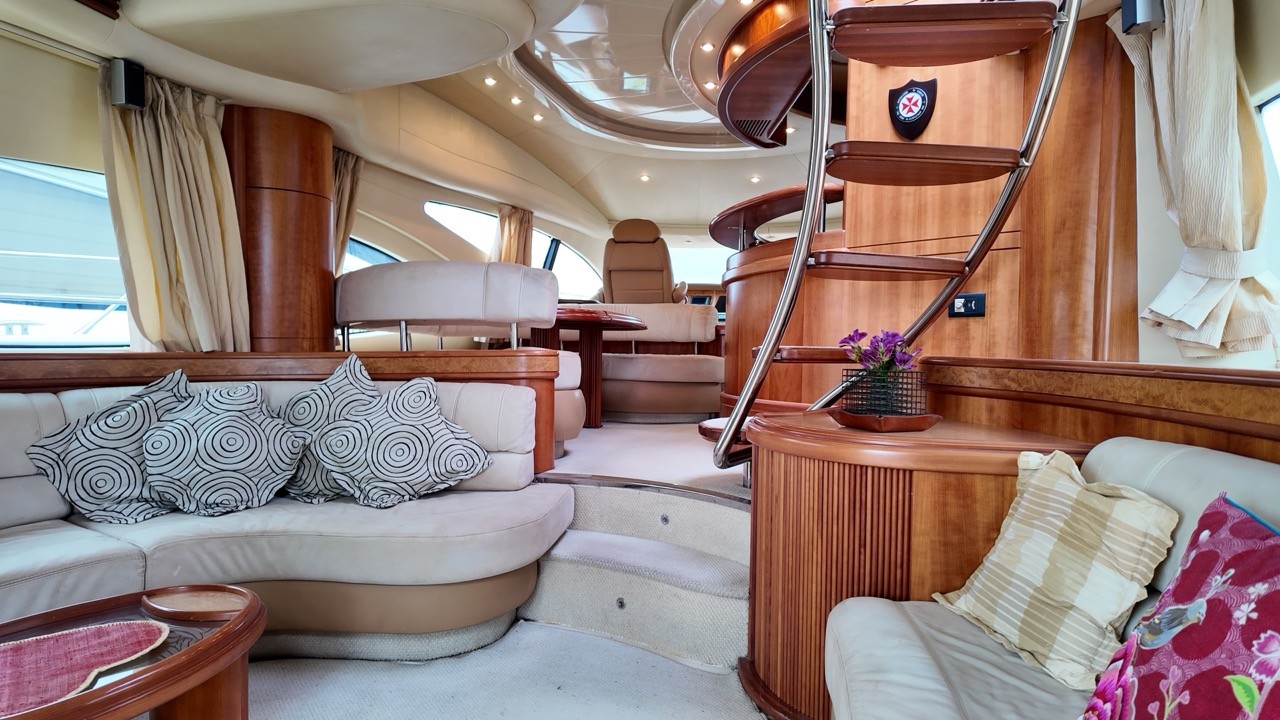Azimut 68 Evolution main saloon interior featuring curved cream leather sofas with patterned cushions, polished mahogany and burl wood cabinetry throughout, circular overhead lighting cove with recessed spots, stainless steel flybridge ladder, and forward helm station beyond