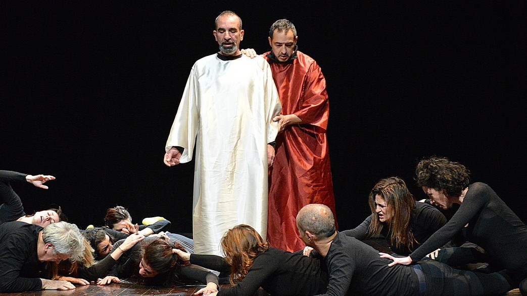 Due attori in lunghi vestiti attraversano il palco pieno di attori distesi