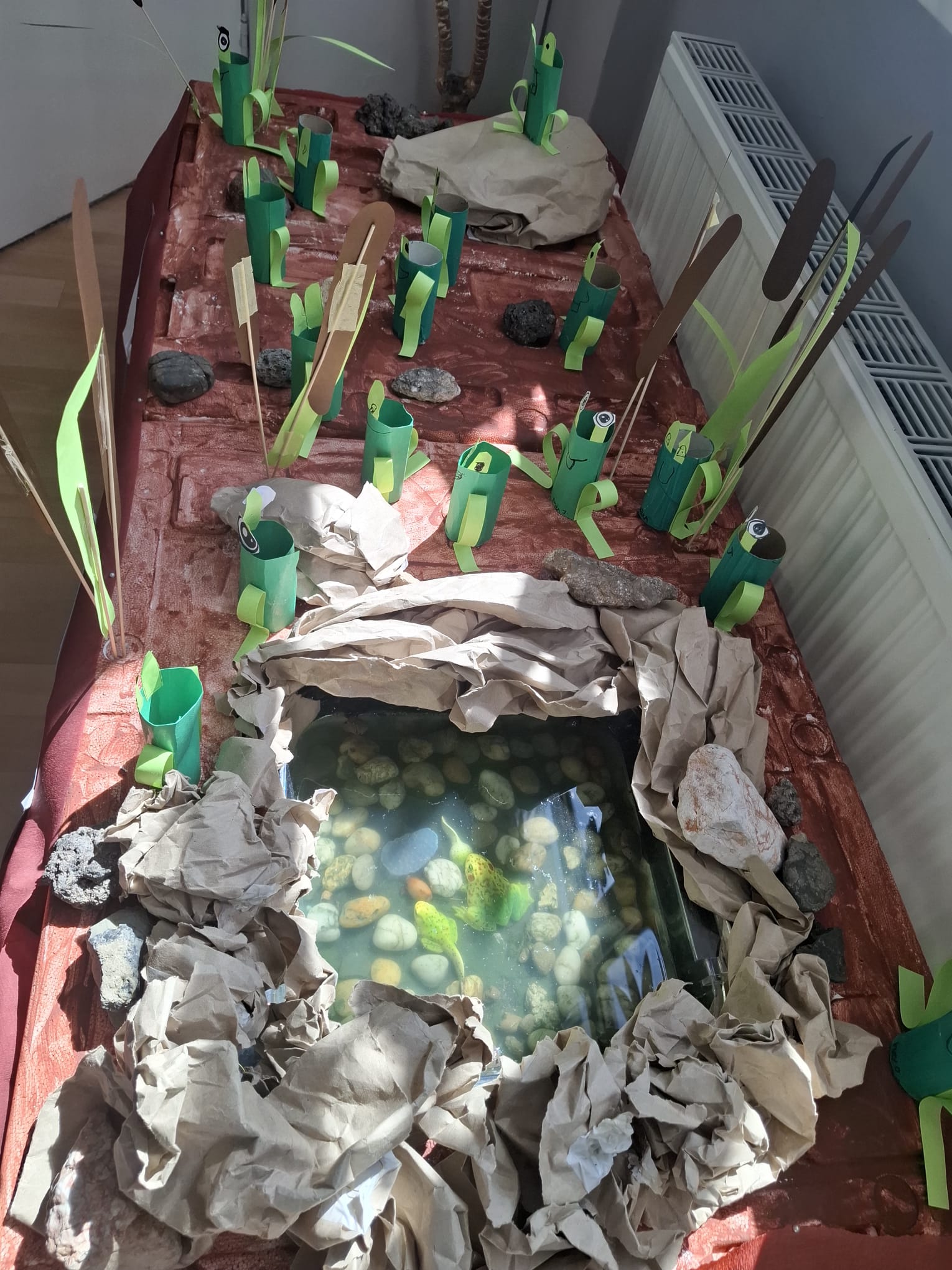 Detailní pohled na model tůně s vodou, obklopený papírovými skalami a zeleným rákosím z papíru.