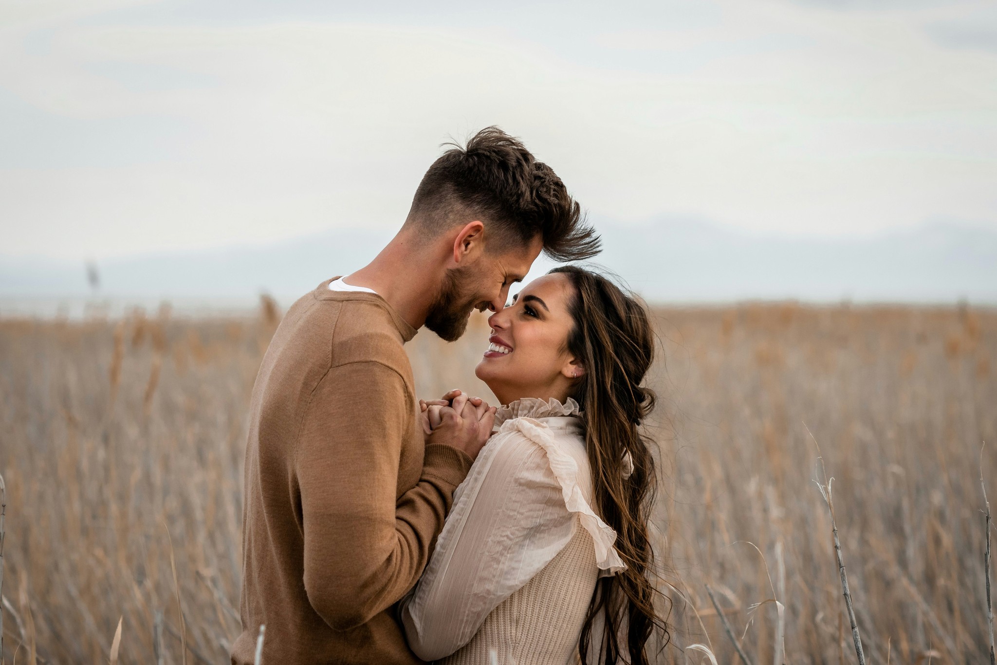 Couple holding hands and smiling nose to nose in a tall grass field with a soft cloudy sky behind them