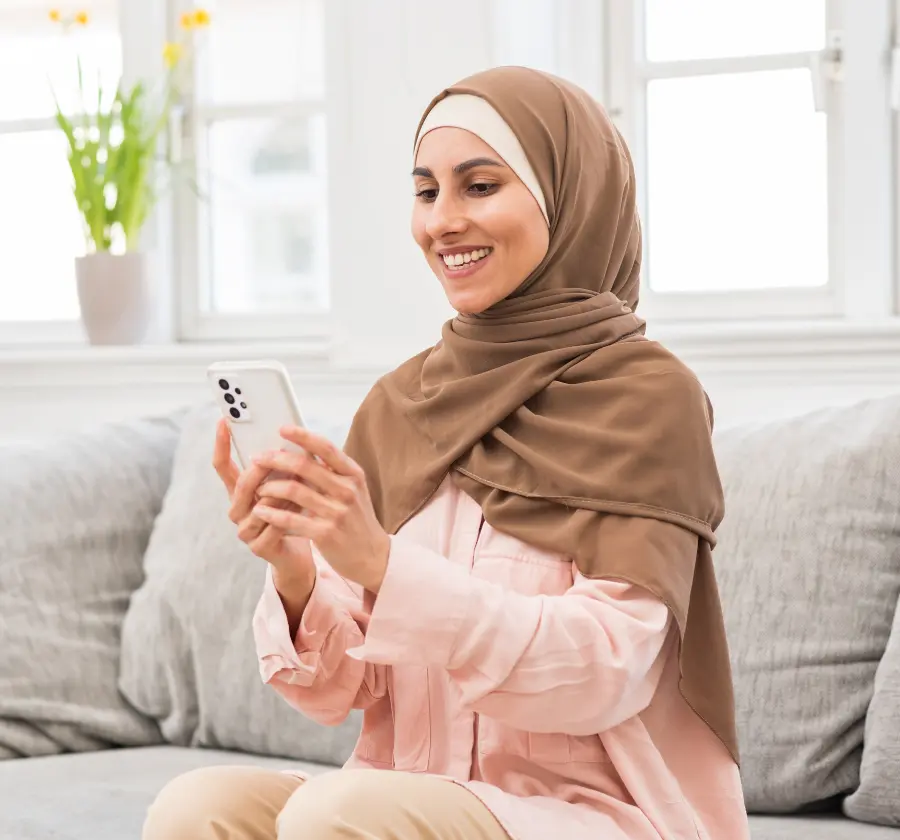 A smiling woman wearing a hijab sits on a couch while using a health app on her smartphone, showcasing digital health solutions for chronic conditions like diabetes in a comfortable, home setting.