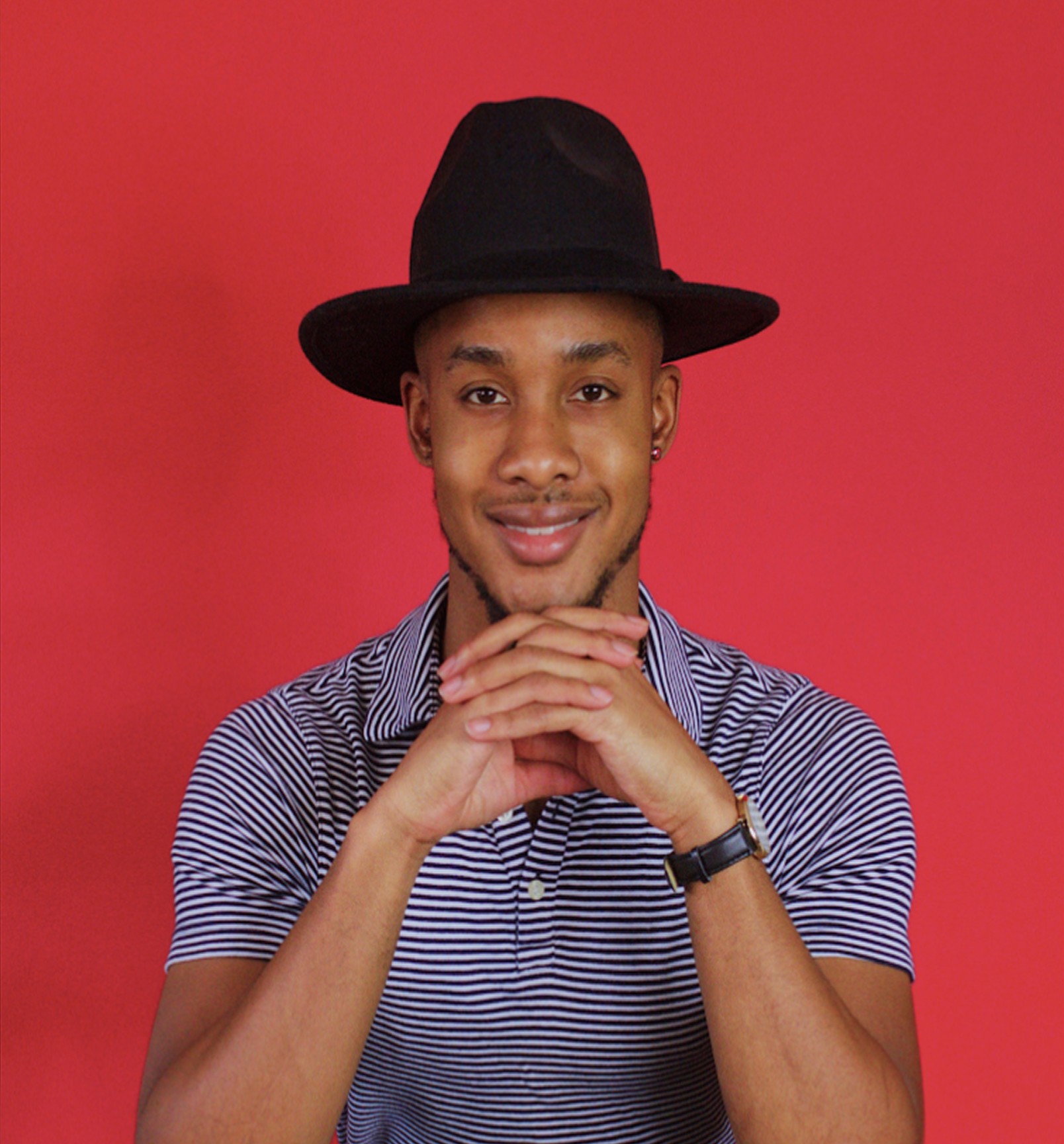 Portrait of Ke’Ron Hall smiling with his hands clasped, wearing a black hat and striped shirt, posed against a solid red background.