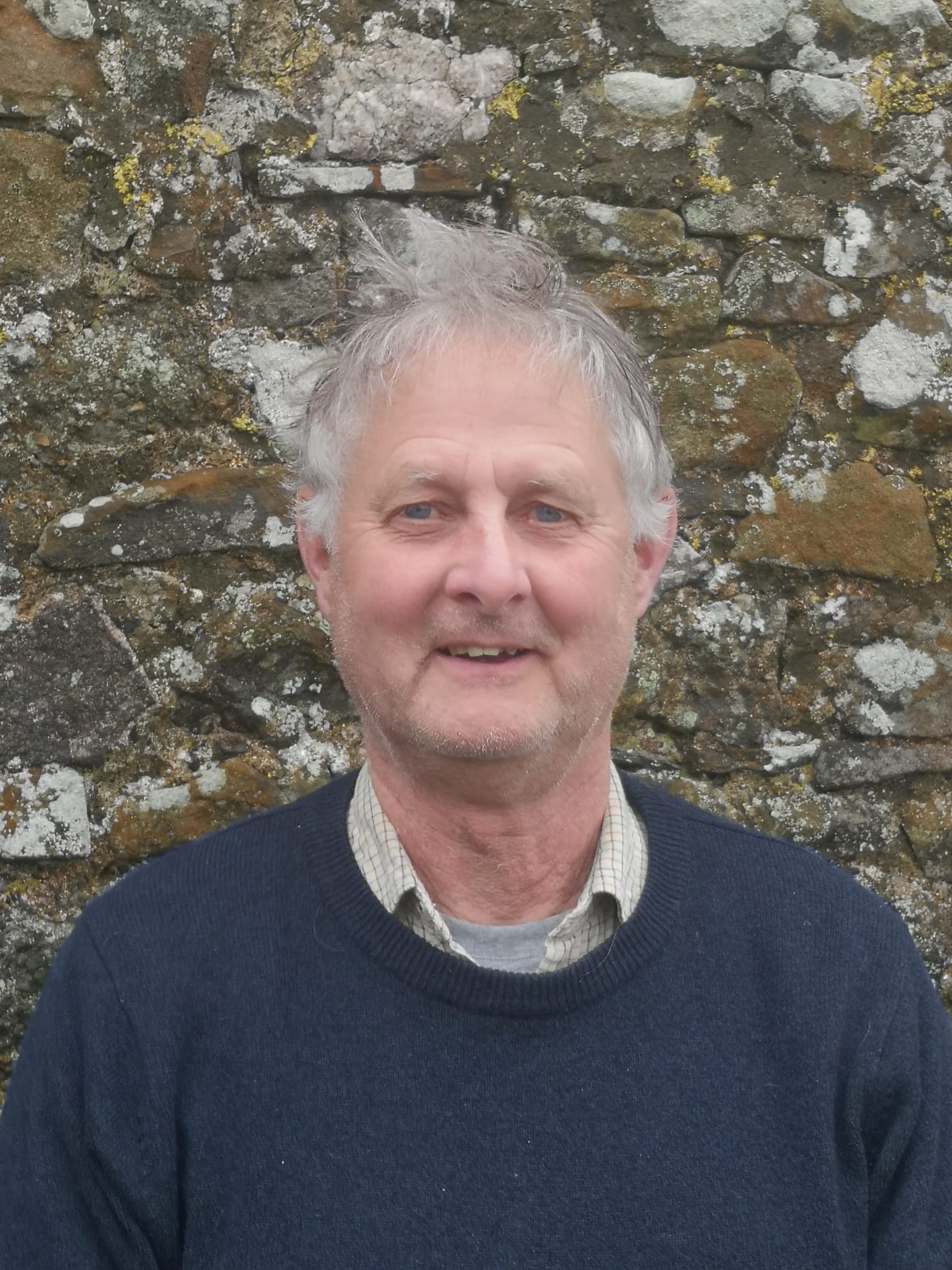 An man standing infront of a brick wall wearing a blue jumper.