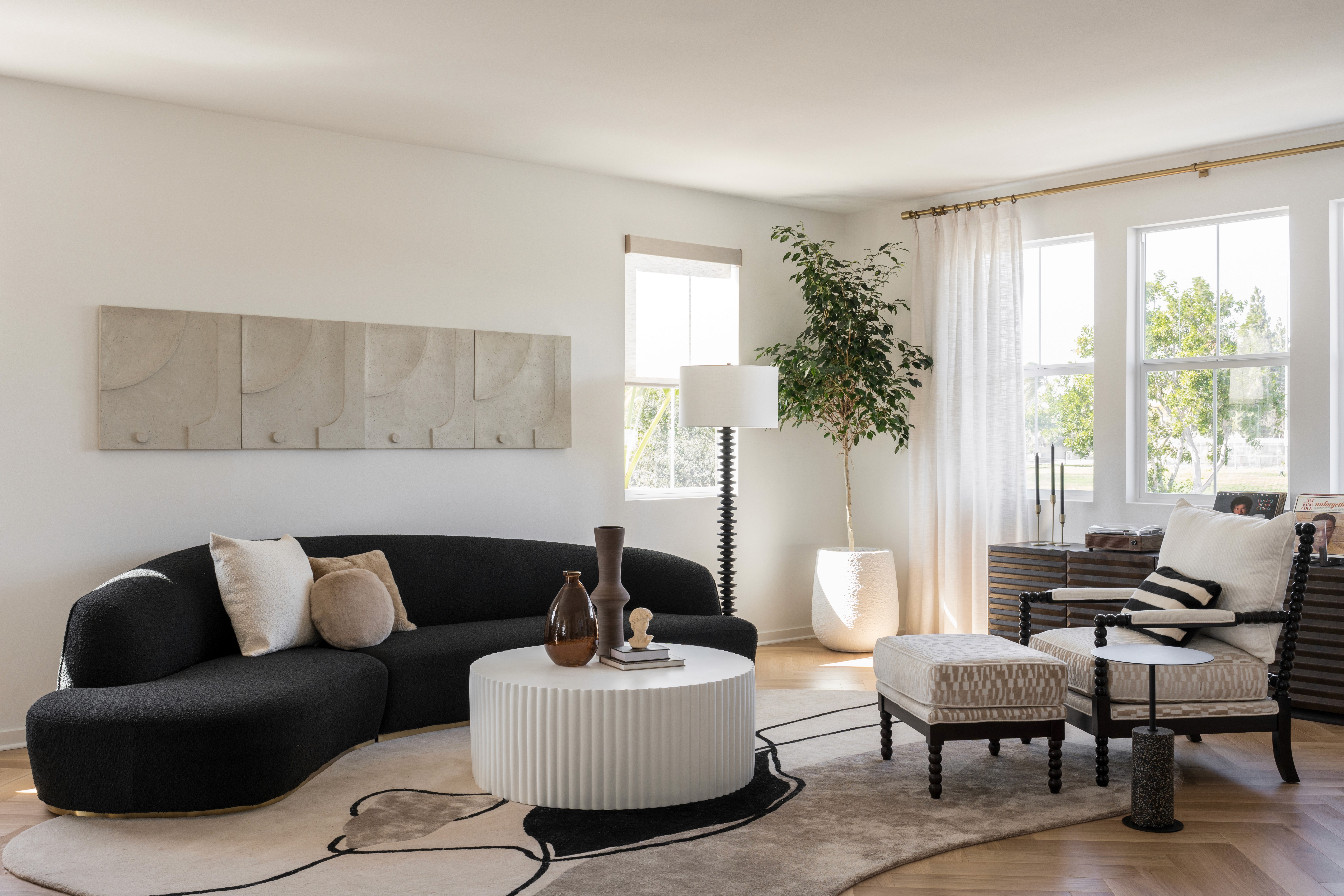 Black kidney bean couch, round, fluted coffee table, large area rug with geometric pattern, modern floor lamp; dark wood console table in background under large windows with floor to ceiling drapes