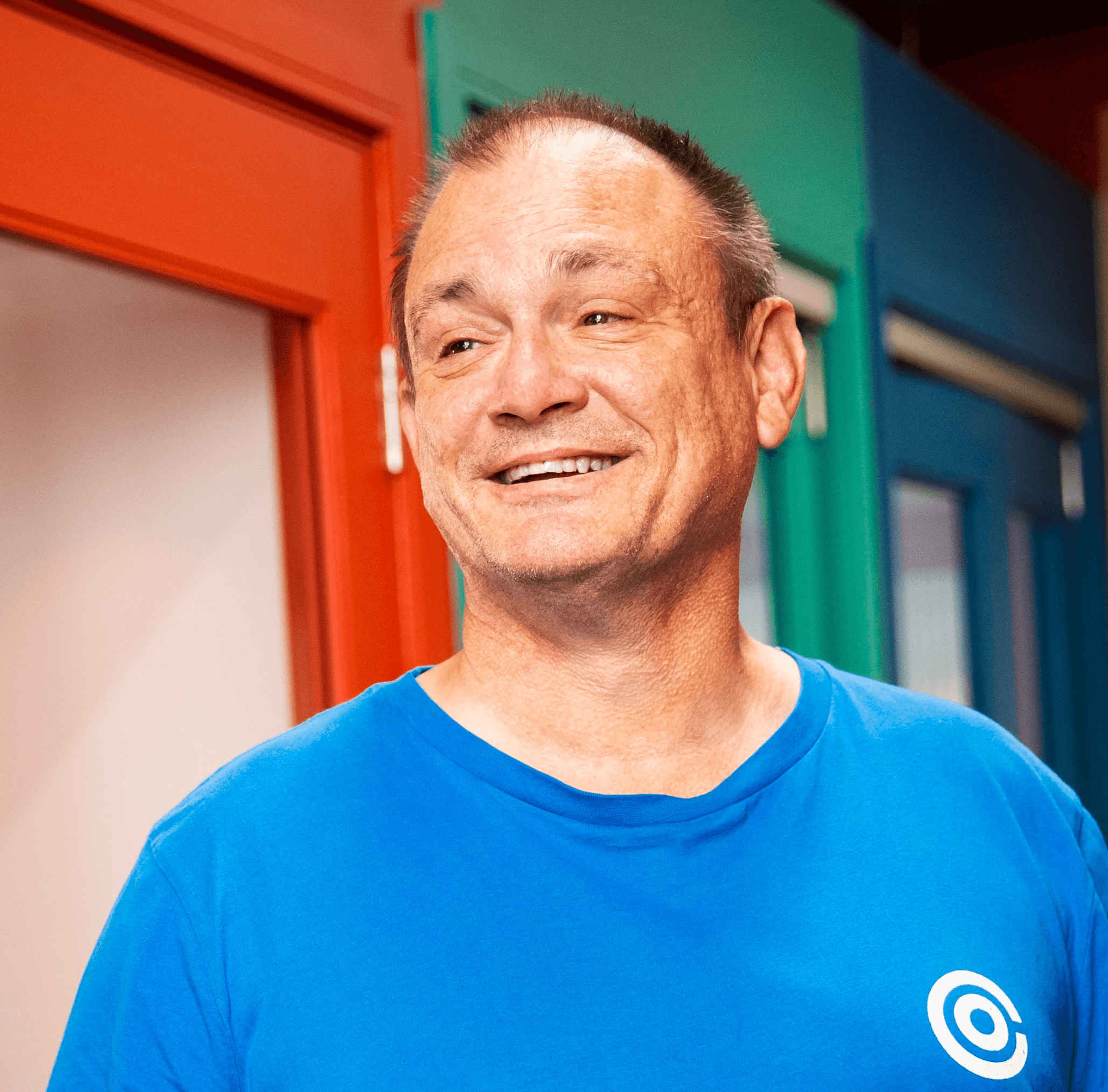 Image of Darren Horobets-Farley, wearing a blue t-shirt with an Opencast logo, standing in a brightly coloured office corridor with red, green and blue doors in the background.