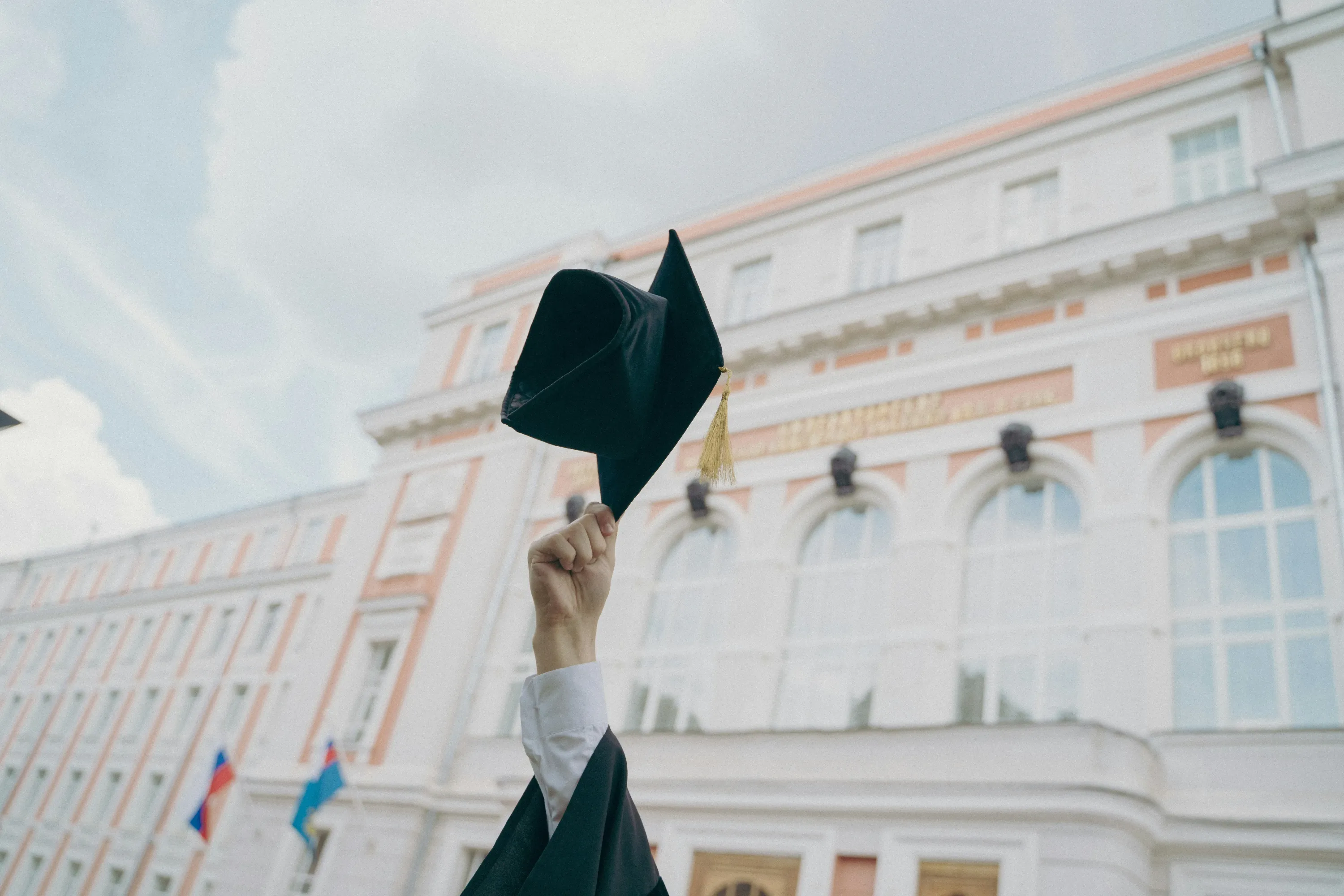 A hand holding a black graduation cap aloft before a large university building