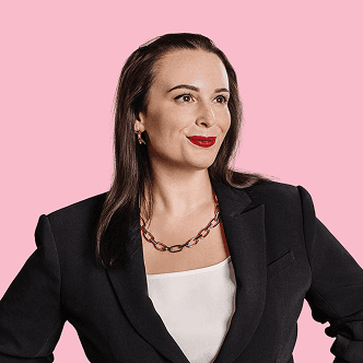 Woman with long dark hair wearing a black blazer and bold necklace, looking to the side against a pink background.