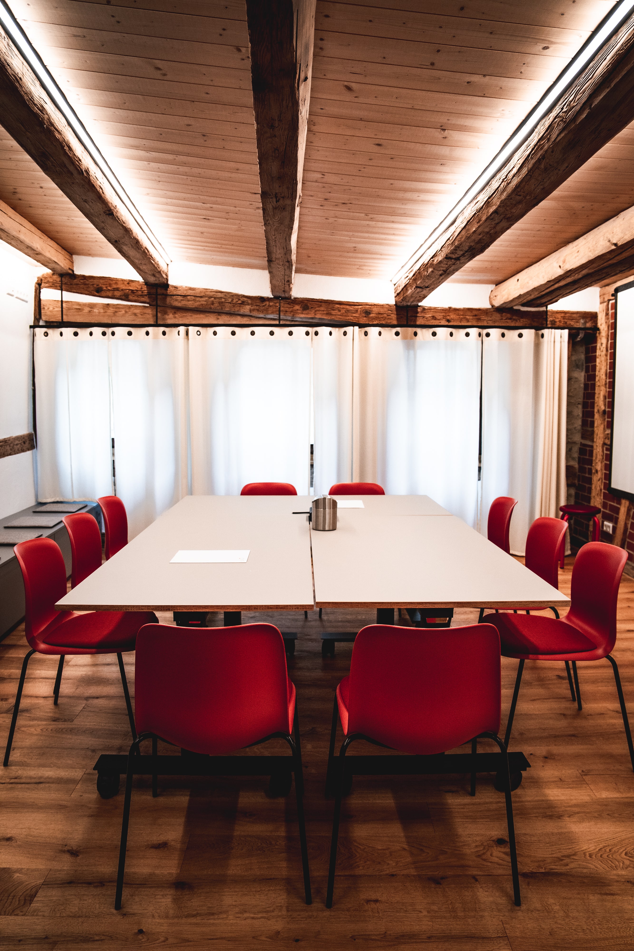 Meeting-Raum mit einem großen Tisch, umgeben von roten Stühlen und hellen, luftigen Fenstern. Elegante Holzbalken über der D