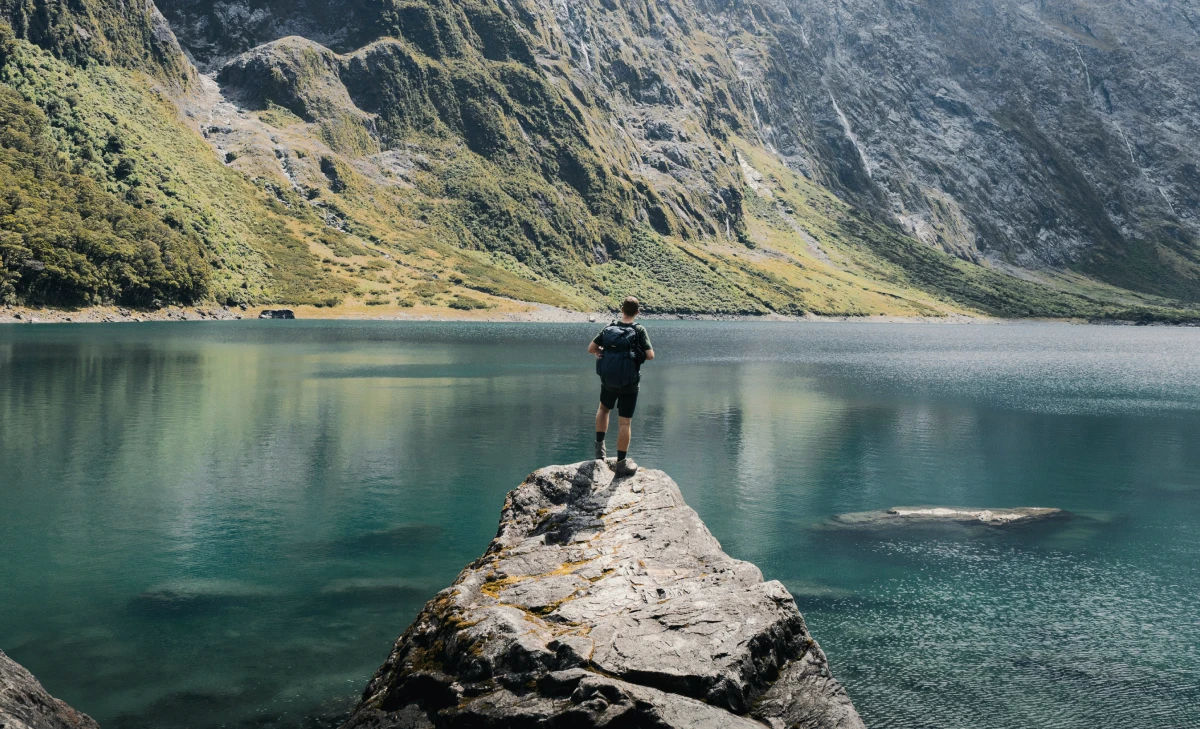 A man stands on a rock, looking at a calm lake surrounded by mountains.