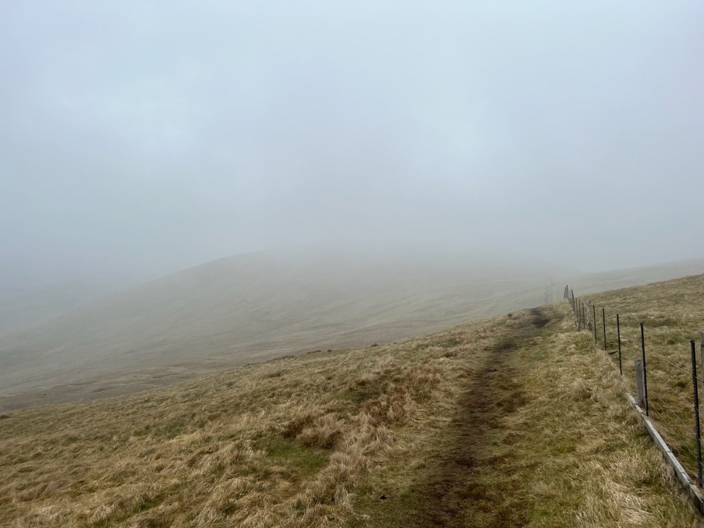 A muddy grass path leading into fog. A fence lines the path on the right. The top half of the image is grey with fog.