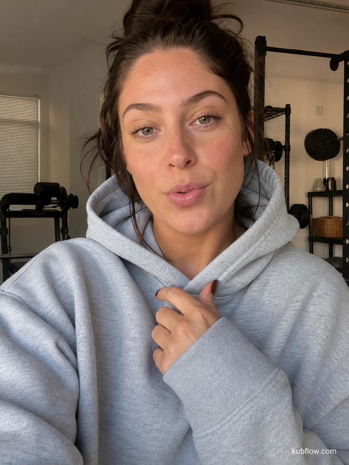 UGC-style photo: beauty creator, in a garage workout corner, trying on a plain hoodie while talking to camera, close vertical talking-head shot.