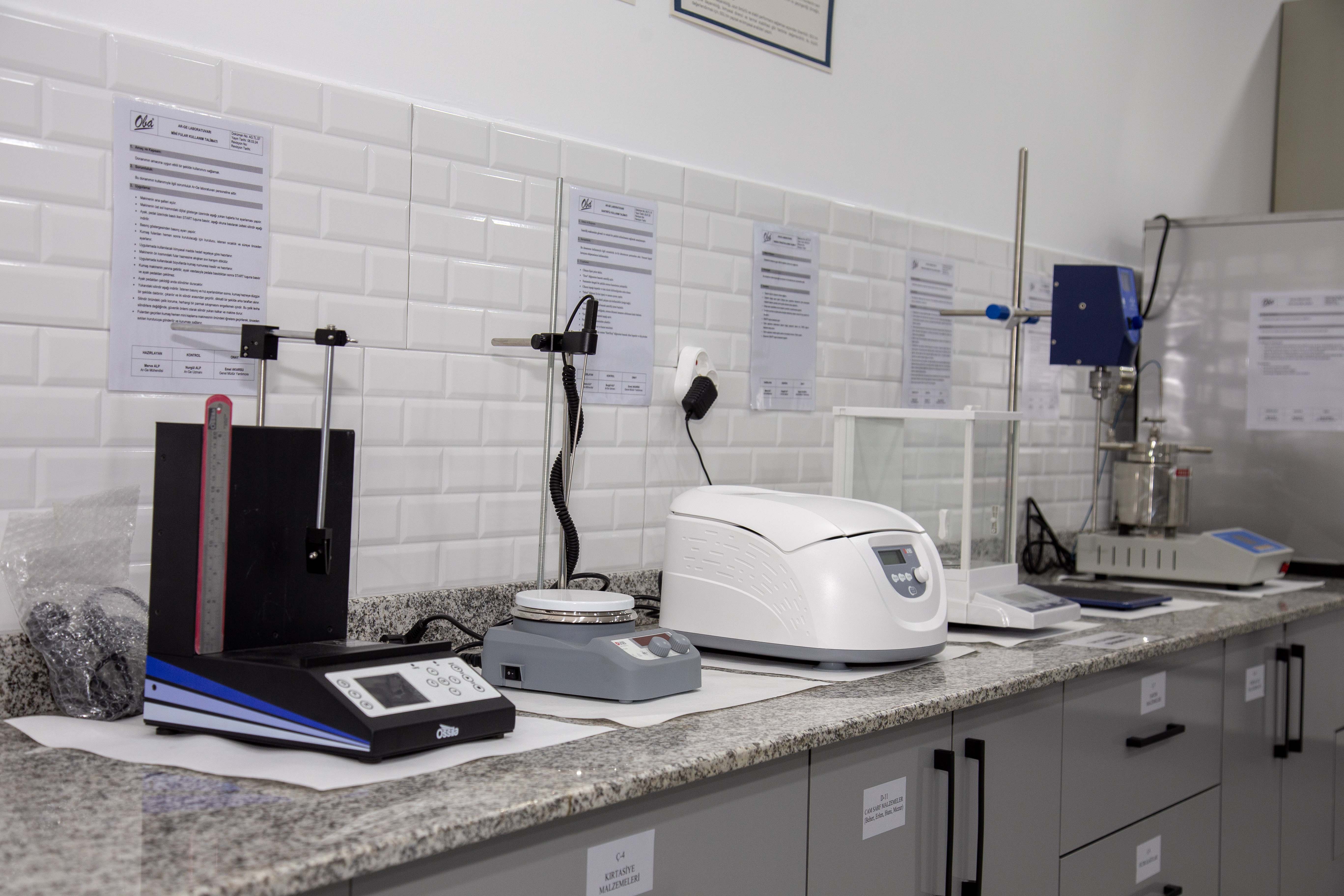 A person sits at a cluttered desk in a dimly lit room, focused on two monitors displaying aerial maps. The walls are covered with various images and notes.