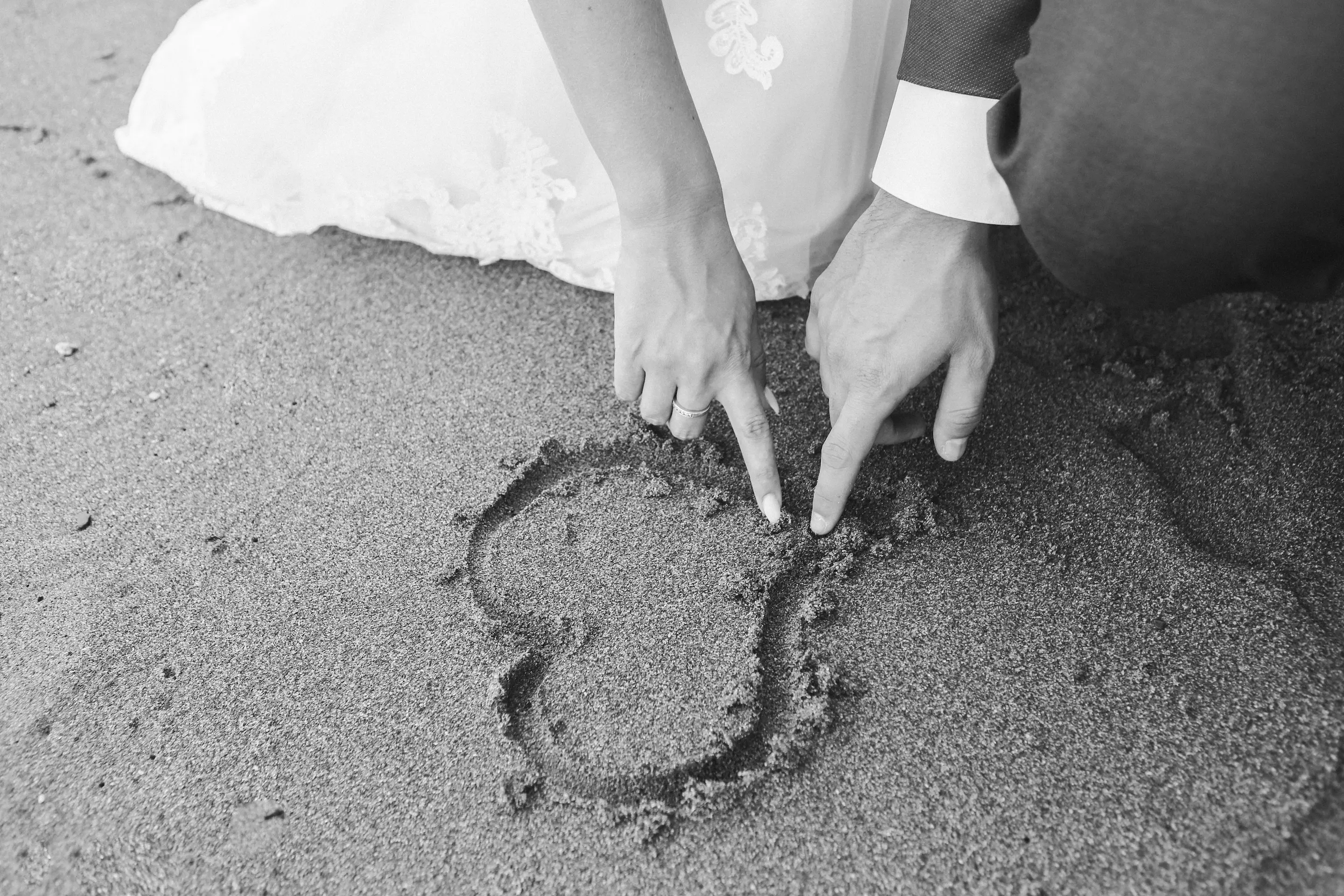 A person in a white dress draws a heart shape in the sand with their fingers.