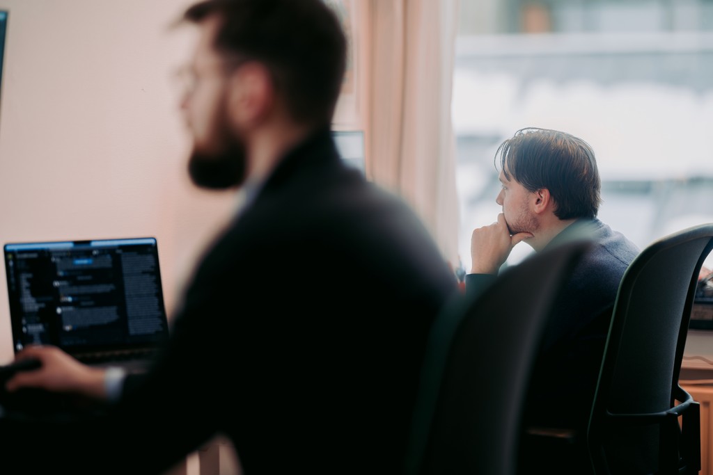 A person in a blue shirt is seated at a desk, thoughtfully gazing outside a window, while another individual in the foreground works on a laptop, creating a focused office environment.