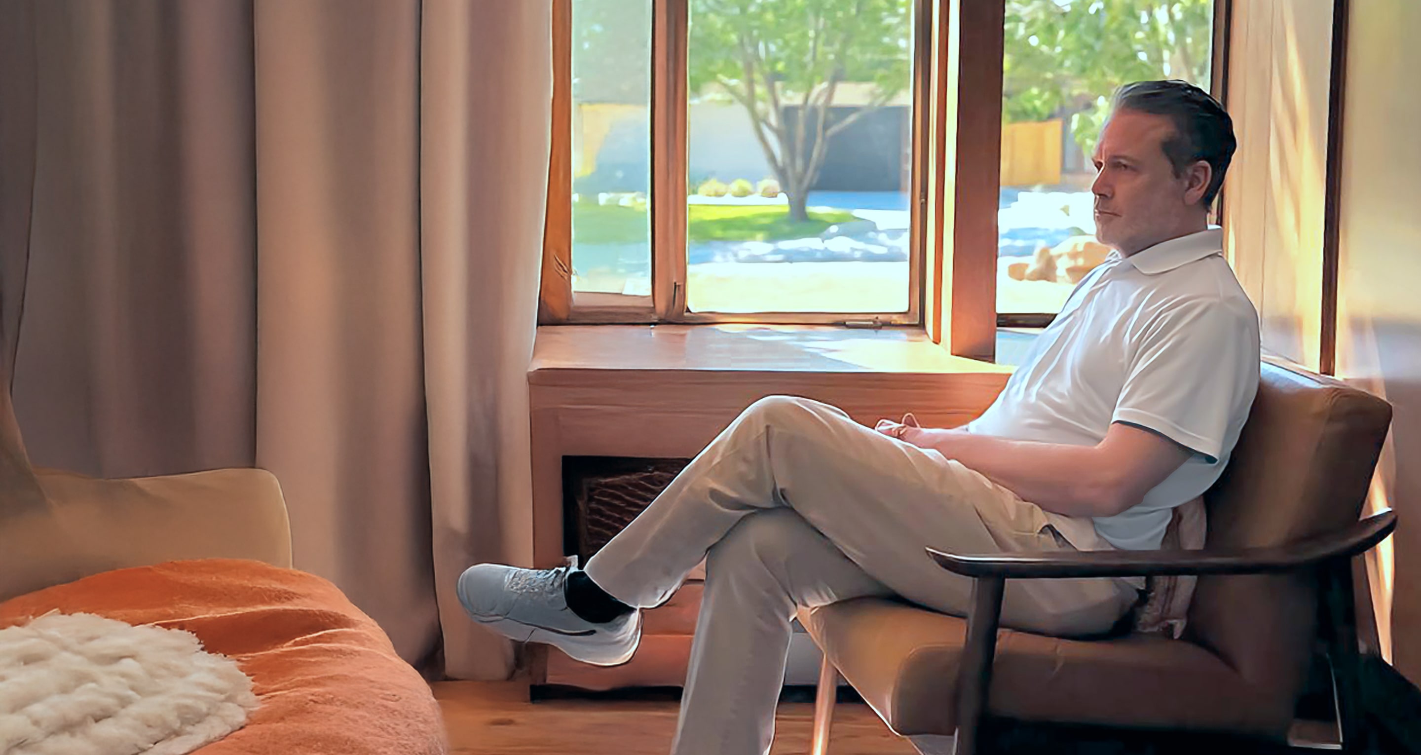Abe Jasinowski (Abe Zemi) sitting in a wooden chair near floor-to-ceiling curtains and a large window overlooking a garden.